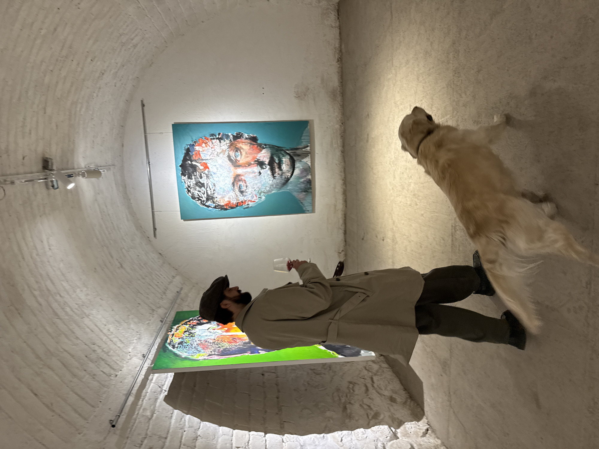 The image shows a man and a dog in a room in Yerevan, Armenia. The room has a curved ceiling and white brick walls. Two abstract paintings are on the wall. The man is wearing a beige trench coat, dark pants, dark shoes, and a dark hat. He is holding a wine glass and looking at the paintings. The dog is a golden retriever.