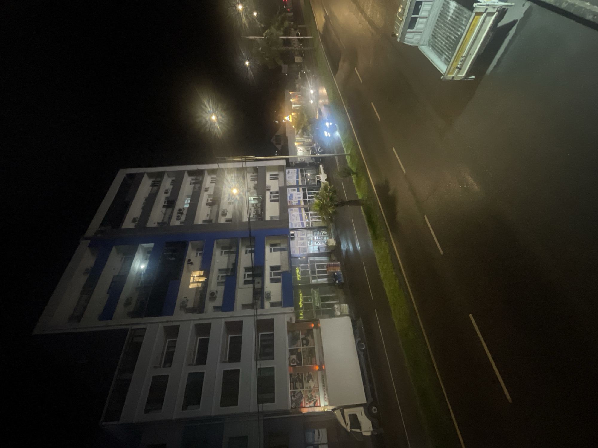 The image depicts a nighttime street scene in Batumi, Georgia, featuring a multi-story building with apartments and commercial spaces on the ground floor, illuminated by interior lights and streetlights. A white box truck is parked on the left side of the street, and several cars are driving on the wet asphalt road. Palm trees line the street, and the overall atmosphere suggests a moderately urban environment. The lighting is predominantly artificial due to the time of day and the presence of street lamps and building lights.