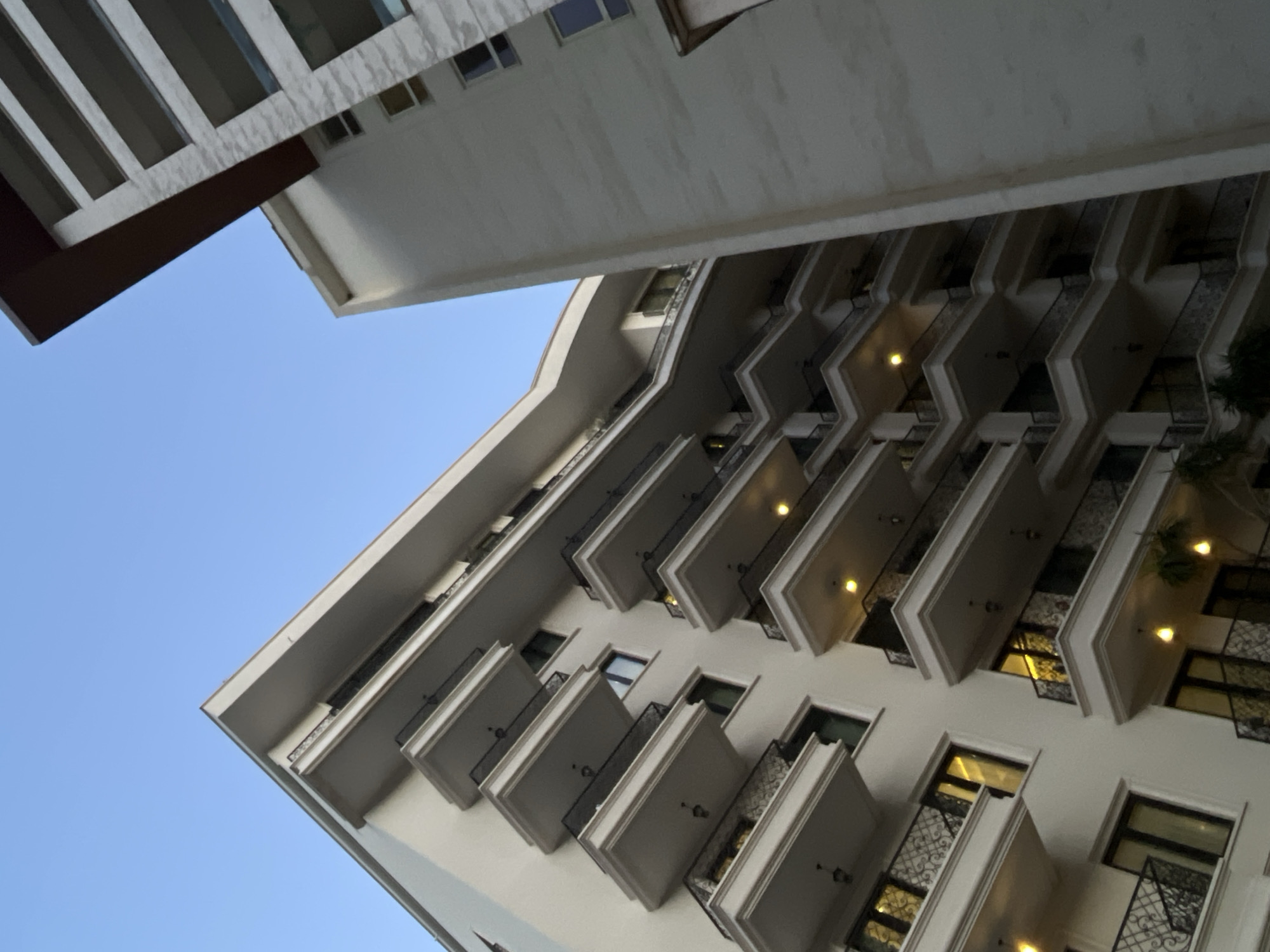 The image shows a low-angle view of a modern apartment building with multiple balconies and windows, set against a clear blue sky. To the right, another building with a textured facade is partially visible. Some of the balconies have ornate railings, and warm yellow lights are illuminated on several levels of the building on the left. The scene is likely captured at dusk or dawn due to the lighting and sky color. The building's architecture suggests it is located in a contemporary urban setting. Based on the architectural style and the context provided, this building is located in Budva, Montenegro.