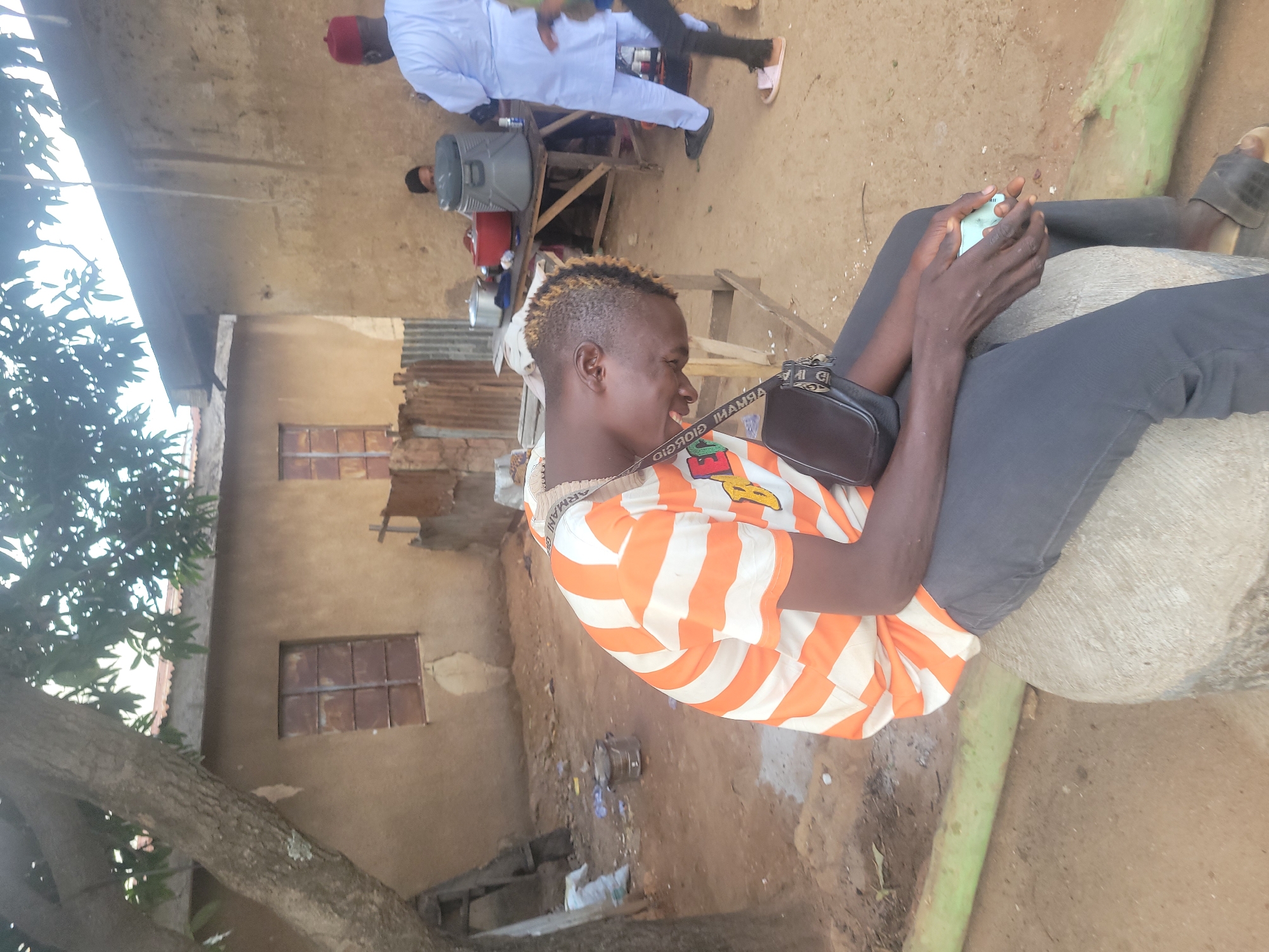 The image depicts an outdoor scene in what appears to be a rural or semi-urban area in Jos, Nigeria. In the foreground, a young person is seated on a concrete cylindrical object, facing down and to the right with their eyes closed. They are wearing a striped orange and white t-shirt, dark grey pants, and black sandals. A small black bag is slung across their chest. Their hands are resting on their lap, and they are holding a light blue object, possibly a mobile phone. To the left of the seated person, a large tree trunk with branches and leaves is visible, casting shadows on the ground.
In the background, there is a light brown, mud-brick building with a corrugated metal roof. The building has two windows with rusted metal bars. Further to the right, another person wearing a light blue traditional outfit and a red cap is standing, facing away from the camera. Behind them, a wooden table is set up with various items, including large food containers and a cooler. Another person is partially visible behind the table. The ground is predominantly dirt and sand. The overall environment suggests a communal or gathering space, possibly a food stall or a residential courtyard.