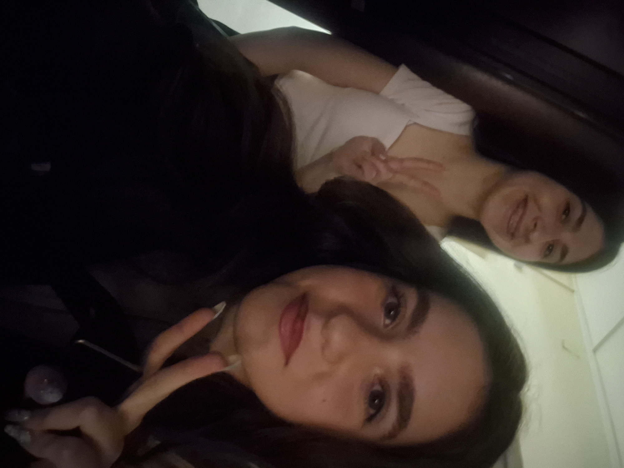 Two women are shown in a selfie. The first woman in the foreground wears a black shirt and red lipstick. The second woman in the background wears a white shirt. Both women are smiling and making peace signs with their hands. The location cannot be confirmed as Pavlikeni, Bulgaria from this image. The background appears to be an interior wall.