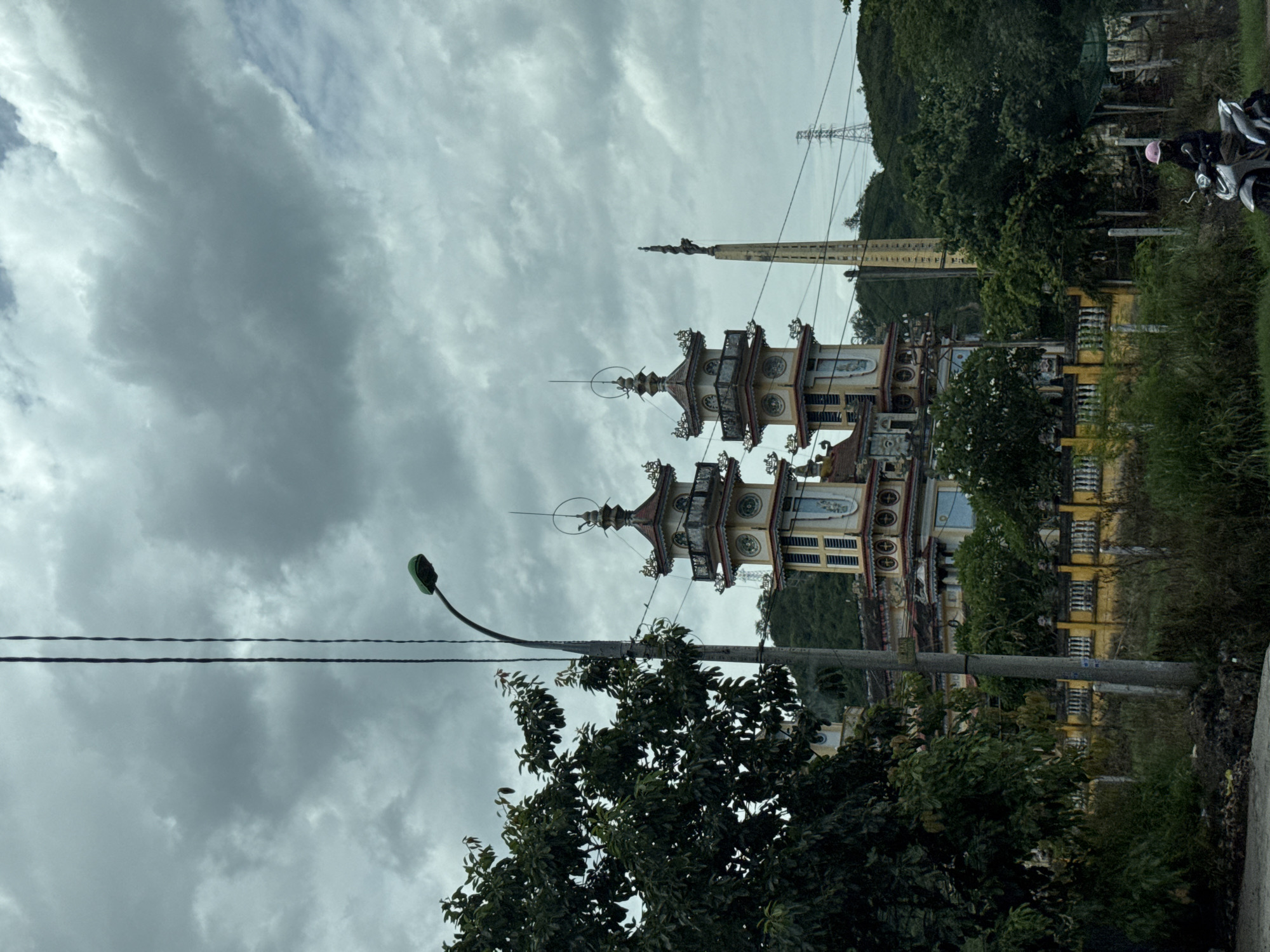 A wide shot captures two ornate, multi-tiered towers with decorative roofs and colorful facades, likely part of a temple complex, set against a backdrop of lush green hills under a cloudy, overcast sky. A tall, slender obelisk topped with a statue stands to the right of the towers. A dark green tree dominates the left foreground, obscuring part of the structures and a streetlamp with a green light. In the middle ground, a yellow fence runs along the perimeter of the complex, with overgrown grass and vegetation in front of it. A man wearing a pink helmet is riding a silver scooter on a dirt path in the lower right corner. Power lines crisscross the scene, with a large transmission tower visible on the hill in the distance. The overall impression is one of an ancient religious site nestled within a tropical landscape, with modern infrastructure subtly present. The scene is likely located in the vicinity of Vĩnh Lương, Vietnam, given the architectural style and the surrounding environment.