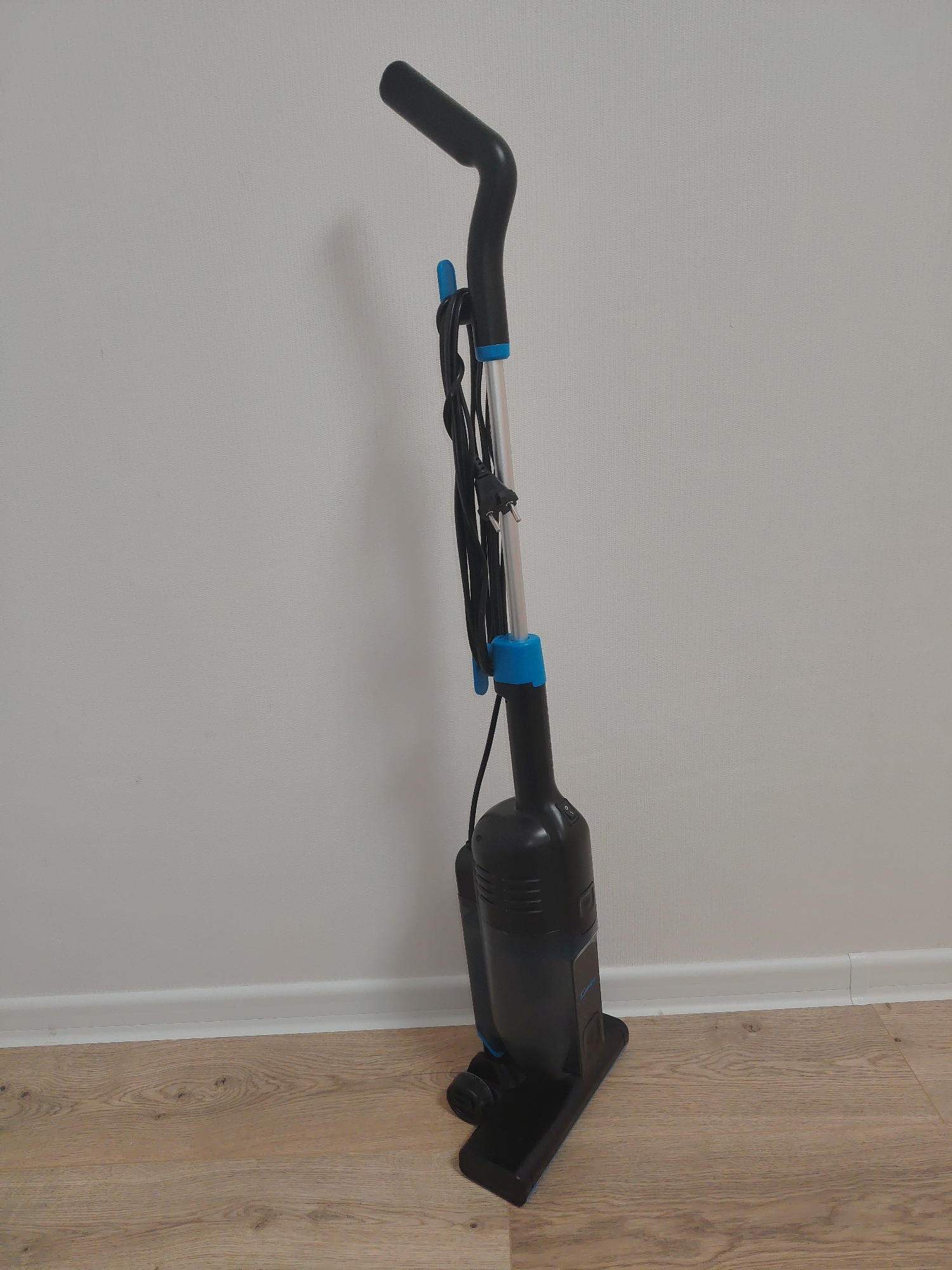 The scene depicts an interior space in Yekaterinburg, Russia. A black and blue stick vacuum cleaner, identified as a 'Candy' brand appliance, stands upright against a light gray textured wall. The cleaner features a curved black handle at the top, a silver metallic shaft, and a main black body with blue accents at the shaft connections. A black power cord, fitted with a European Type F (Schuko) plug, is loosely wrapped around the upper section of the shaft. A power switch is visible on the upper rear of the main body. The base of the appliance is a flat, rectangular black head resting on a light-toned wooden laminate floor. A white skirting board forms the junction between the wall and the floor. The space is evenly lit, and the surfaces appear clean.