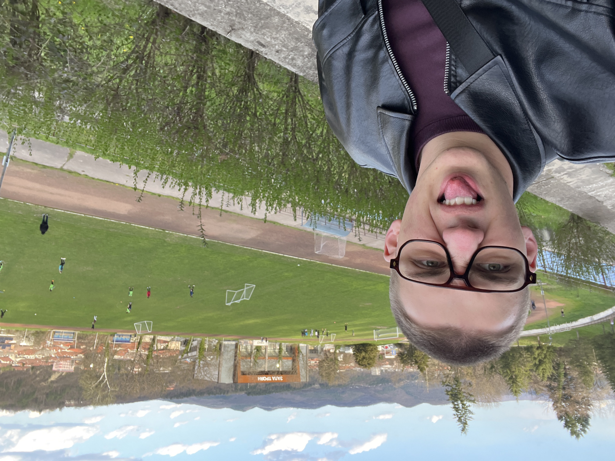 A selfie-style image features a person with short, light hair, wearing glasses and a black leather jacket. The person is sticking their tongue out slightly. In the background, a sports stadium with a green field is visible, where multiple individuals are playing sports. Beyond the stadium, a cityscape is apparent, set against a backdrop of mountains and a partly cloudy sky. "ЗАЛА ТРОЯН" is written above the building facing the field. This is most likely the stadium in Troyan, Bulgaria.