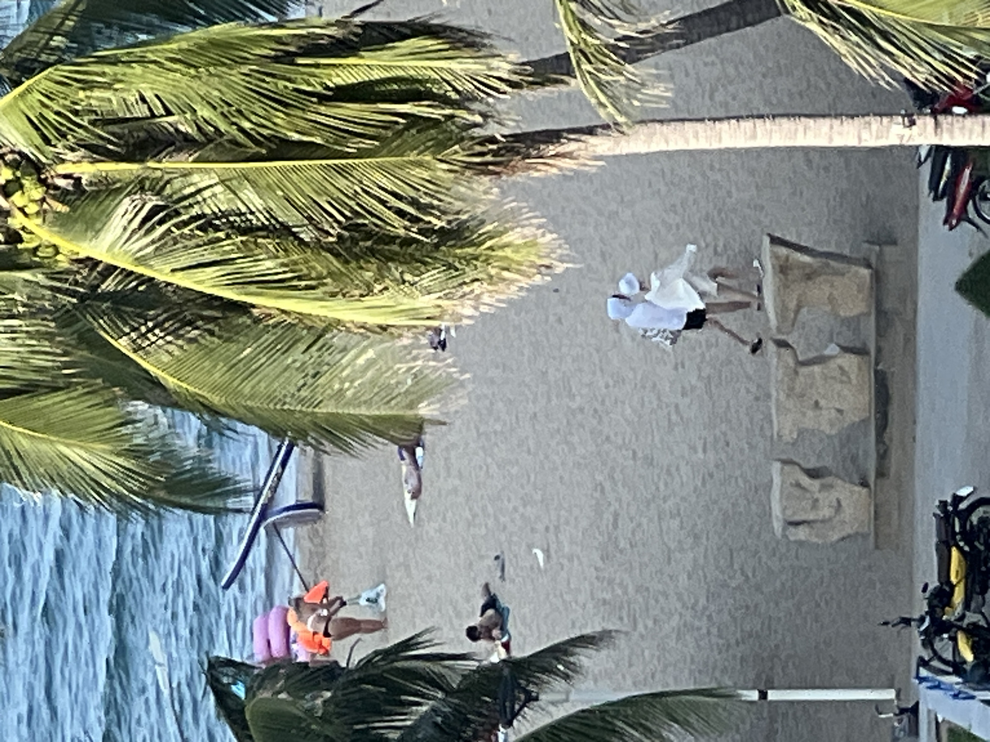 This is an outdoor, eye-level shot of a beach in Nha Trang, Vietnam, featuring palm trees in the foreground partially obscuring the view. The ocean is visible in the background, with several people on the sand near the water. One person, wearing an orange life vest and black bikini bottoms, is near a paddleboard. Further up the beach, two individuals in white clothing are walking. In the middle ground, two people are sunbathing on the sand. Closer to the foreground, a yellow and black motorcycle is parked on a paved area. In the background, large rock sculptures are visible. The image is slightly grainy and taken from a distance.