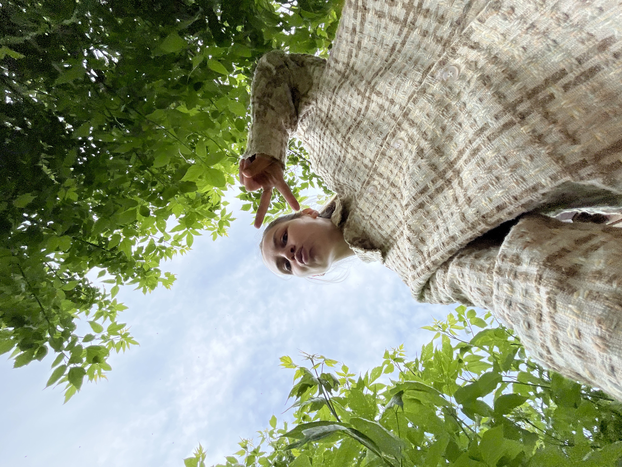 An upward angle photograph captures a person from the chin up, holding their hand in a peace sign. The person appears to be standing outdoors surrounded by trees. The sky is visible through the branches. The person is wearing a patterned beige and brown colored jacket. The location is not discernible from the photograph.