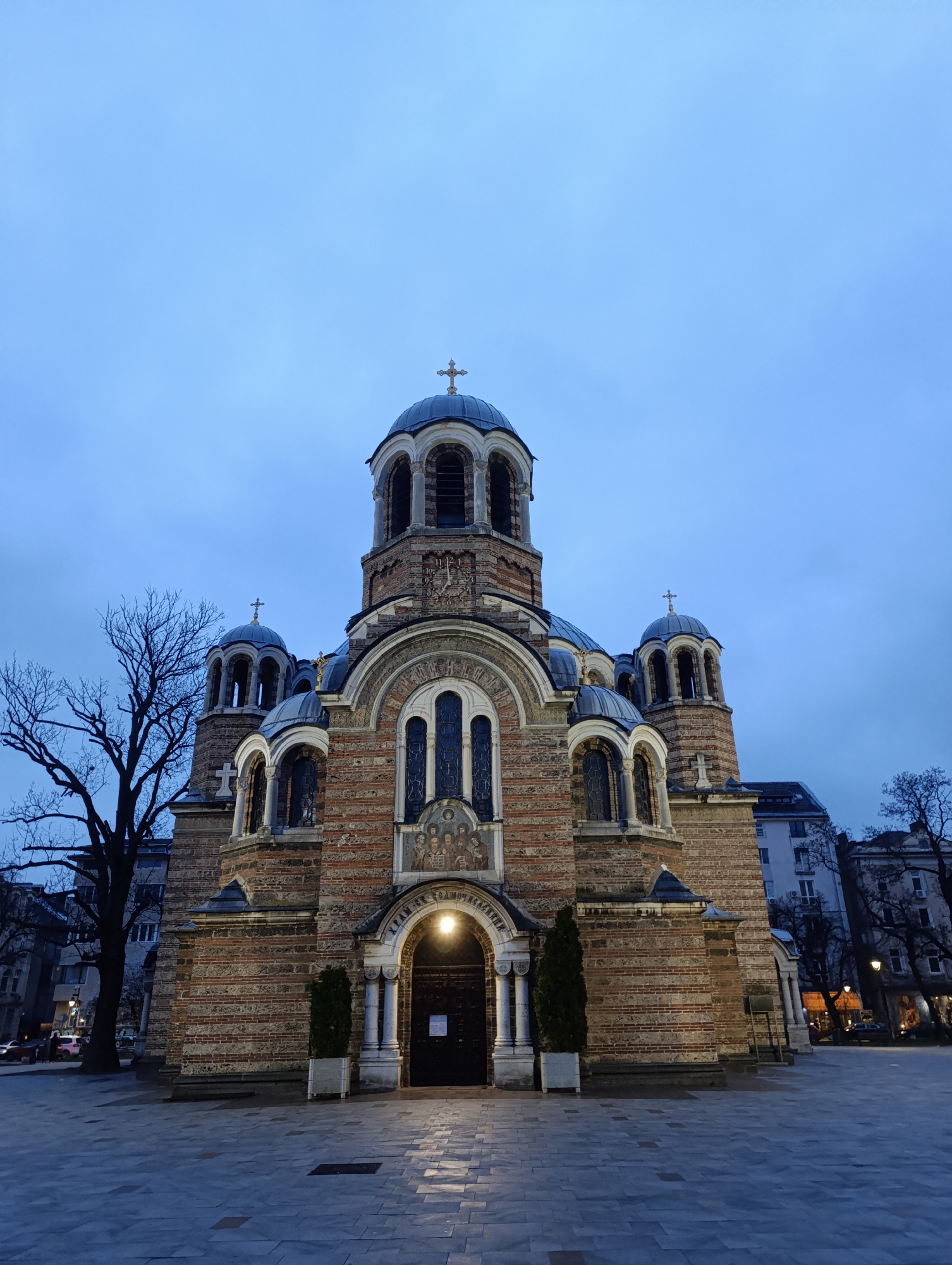 The image captures the exterior of the Church of Sveti Sedmochislenitsi, a Bulgarian Orthodox church located in Sofia, Bulgaria. The church is a multi-domed structure with a brick and stone facade. The main entrance is centrally positioned with white pillars and two small evergreen trees planted in square pots on either side. Above the entrance is an inscription in Cyrillic. The sky is a dusky blue, indicating either dusk or dawn. To the left of the church is a bare tree, and there are other buildings visible in the background. The foreground consists of a paved area, likely a pedestrian square. There is no indication of human activity, and no people are visible in the image.