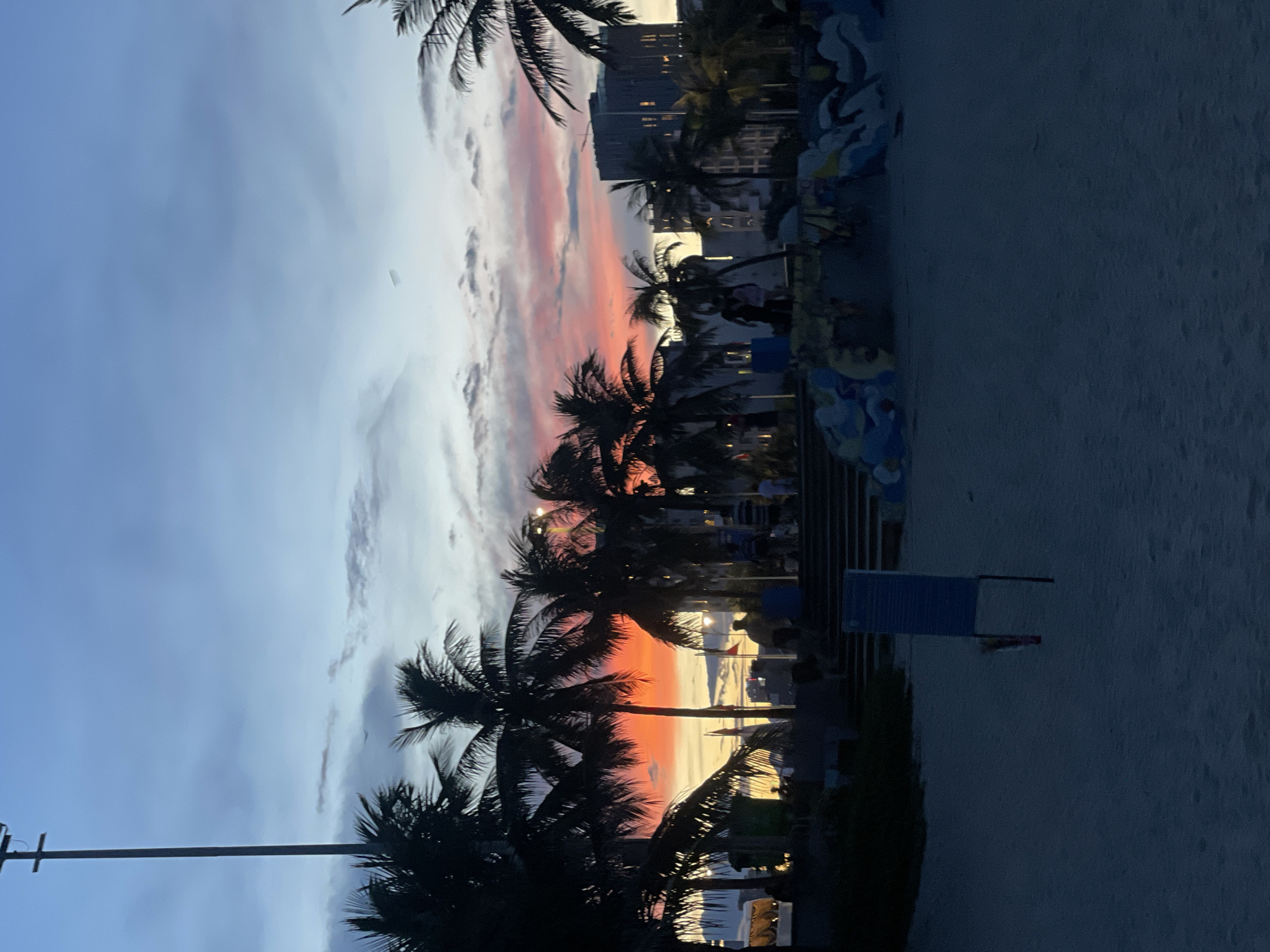 A sunset scene unfolds over a beach area in Da Nang, Vietnam, with a vibrant sky painted in hues of orange, pink, and blue, transitioning into a twilight sky. Silhouetted palm trees dominate the foreground and midground, their fronds fanned out against the colorful sky. In the background, the city skyline emerges, featuring buildings and a prominent street light on the left side. A group of people are visible, some walking and others gathered near stairs and a landscaped area, all rendered in silhouette due to the low light conditions. The ground is covered in sand, suggesting a public beach or promenade. A blue signpost stands in the sand in the foreground.