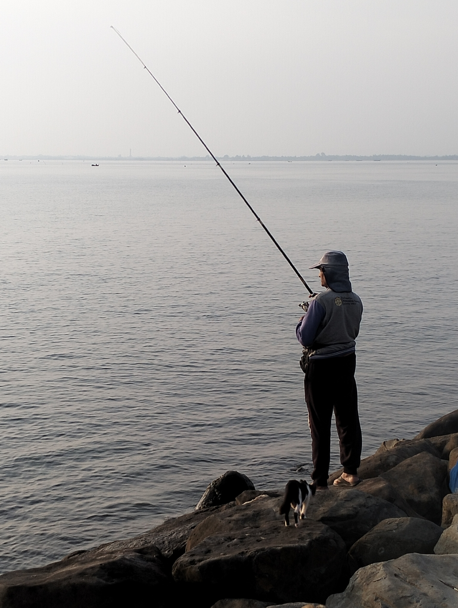 A person stands on a rocky shore fishing. They wear dark pants, a light gray hooded jacket, and a hat. A fishing rod is held in their hands, extended high and at an angle over a body of water. A small black and white cat stands on the rocks near the person's feet. The water is relatively calm with subtle waves. In the distance, a faint skyline and a few small boats can be seen. The scene appears to be from a coastal area. This is in the city of Lhokseumawe, Indonesia.