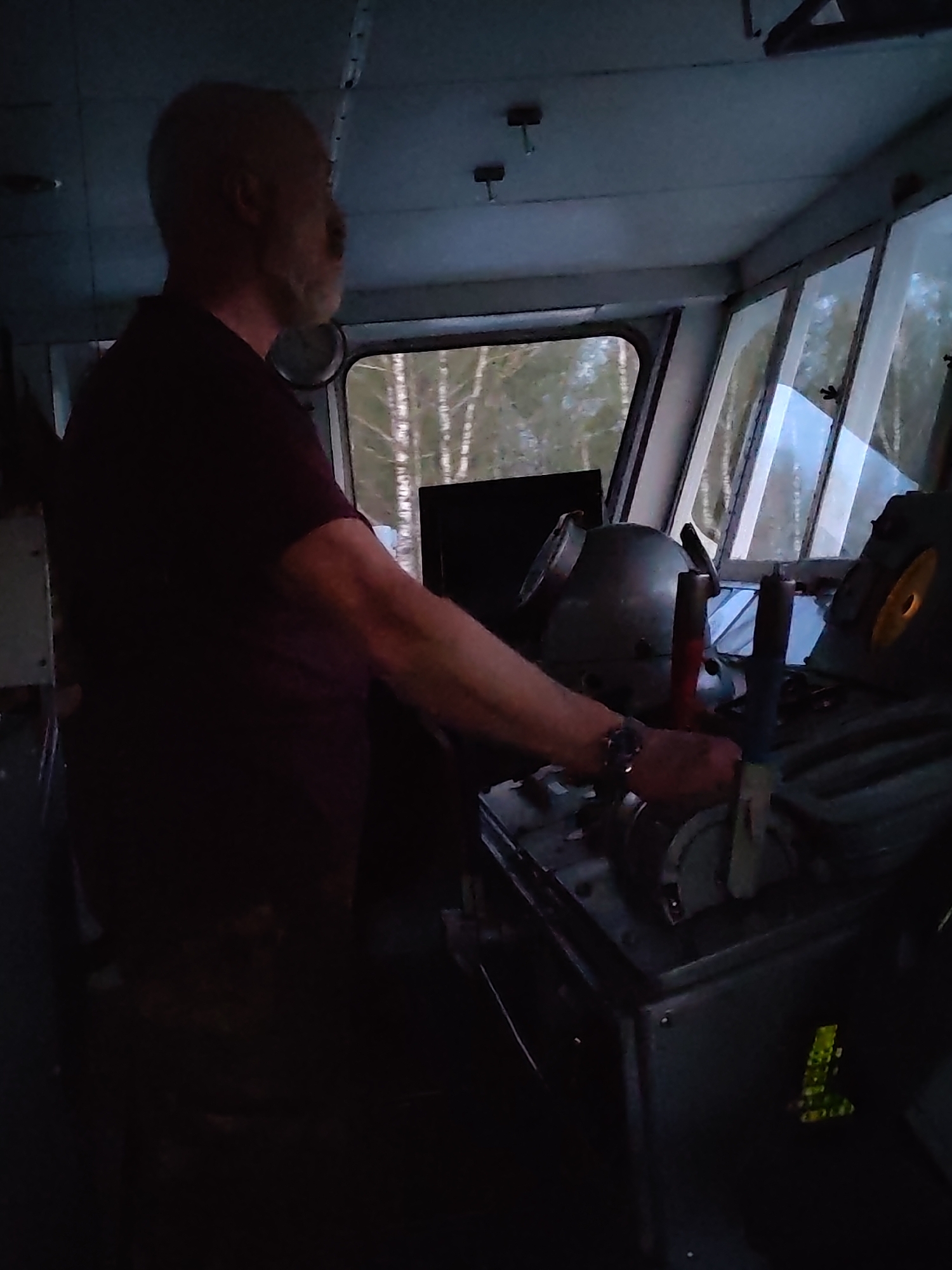 A man is standing in what appears to be the bridge of a boat or ship. The man is bald with a gray beard, wearing a dark shirt and appears to be operating the controls of the vessel. The bridge has large windows that allow a view of a forest of birch trees outside. The location is unknown.