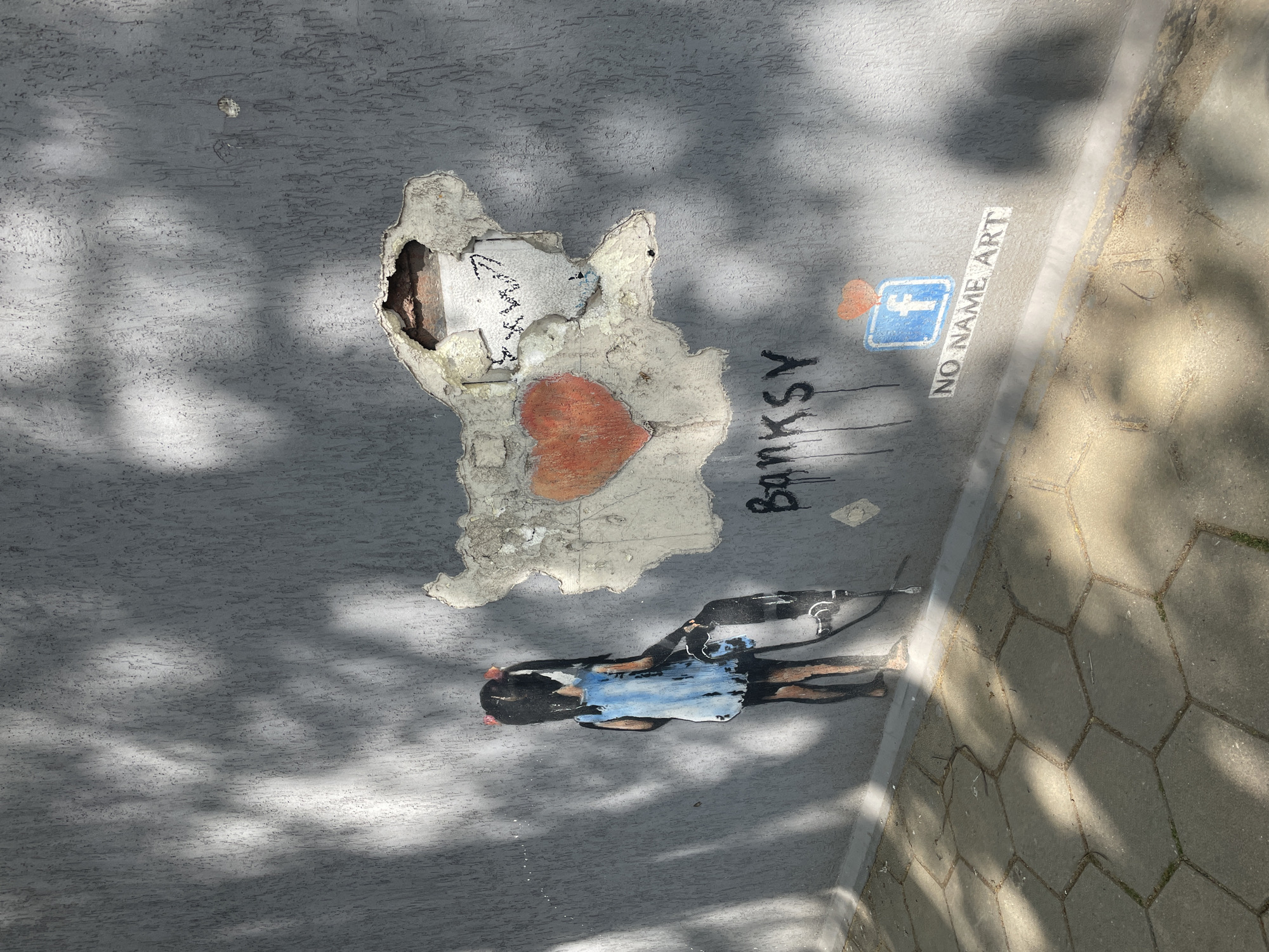 This media file captures a piece of street art or graffiti on a textured grey surface, likely a wall or ground, in Plovdiv, Bulgaria. The scene is viewed from above under bright sunlight, which casts dappled shadows across the surface.
The central artwork features a stencil of a young girl wearing a blue dress, depicted as if pulling a wheeled object or cart. To the right of the girl, the word "BANKSY" is spray-painted in black capital letters, with drips running downwards.
Above the "BANKSY" text, an irregularly shaped section of the grey surface has peeled away, exposing a rougher, lighter-colored underlayer. A red heart is painted on this exposed material. Within the damaged area, faint, scribbled markings or text are barely visible, and a deeper cavity reveals reddish-brown material inside. A smaller, circular hole is also present in the grey surface further up.
Near the "BANKSY" text, a blue square containing a white lowercase 'f' symbol (resembling the Facebook logo) is depicted. Next to this logo, the phrase "NO NAME ART" is written vertically in white letters. The right side of the image features a boundary leading to a sidewalk paved with hexagonal concrete blocks. The clear, defined shadows indicate it is daytime.