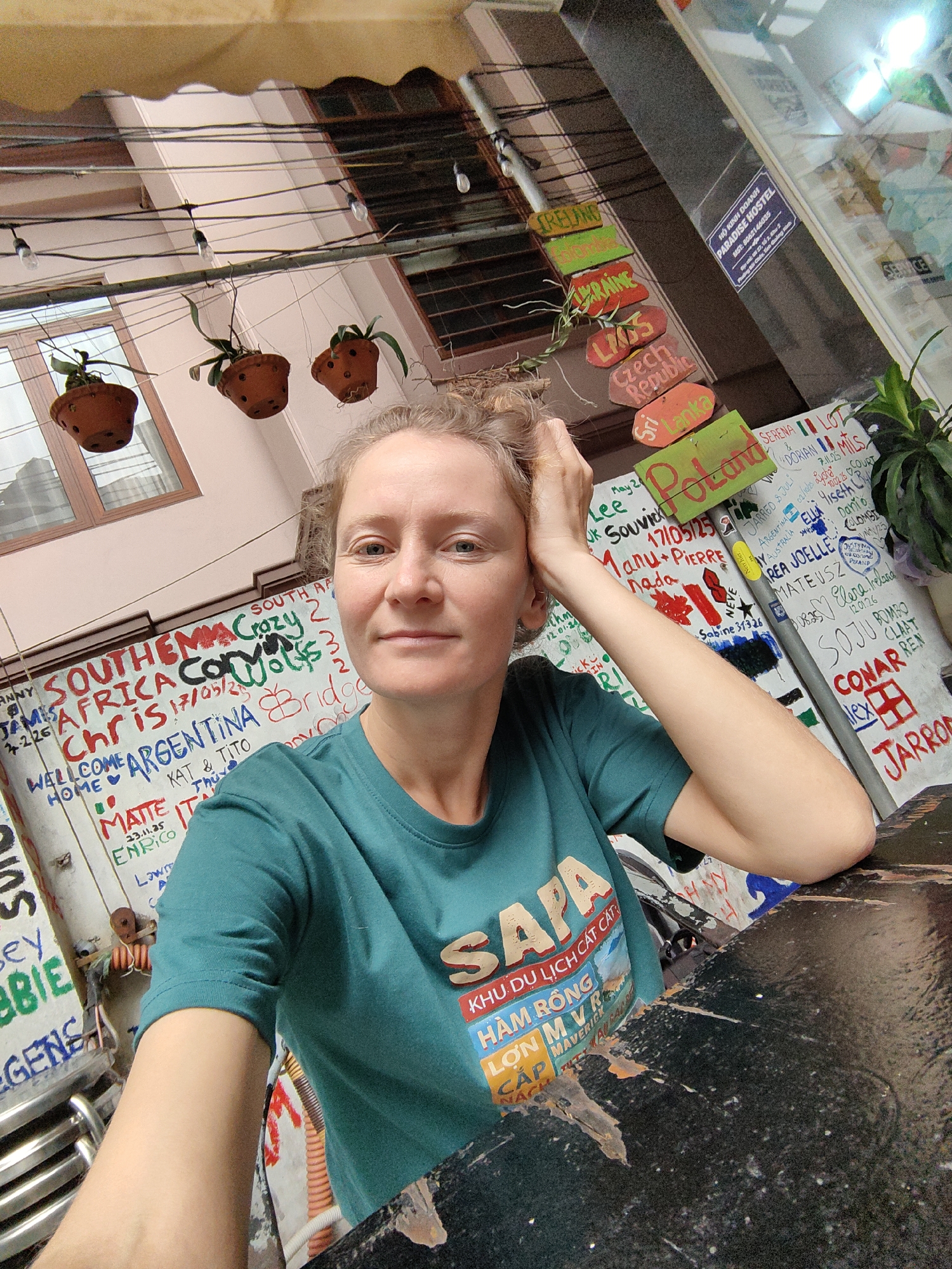 A fair-skinned woman with light brown hair is centered in a selfie-style photograph. She is wearing a teal t-shirt with a print that includes the word "SAPA" and Vietnamese text, partially obscured. Her right hand is resting on her head.
The background is cluttered and vibrant. A wall covered in signatures, dates, and place names like "South Africa," "Argentina," and "Poland" acts as a backdrop. A wooden signpost with country names including "Ireland," "Ukraine," "Laos," "Czech Republic," and "Sri Lanka" hangs overhead. Small, hanging potted plants are visible above.
A sign indicating "Paradise Hostel" (HỘ KINH DOANH PARADISE HOSTEL) with the address and MST number, places the location in Bai Chay Ward, Vietnam. A black, distressed table occupies the foreground. Electrical wiring and various objects add to the chaotic outdoor setting.