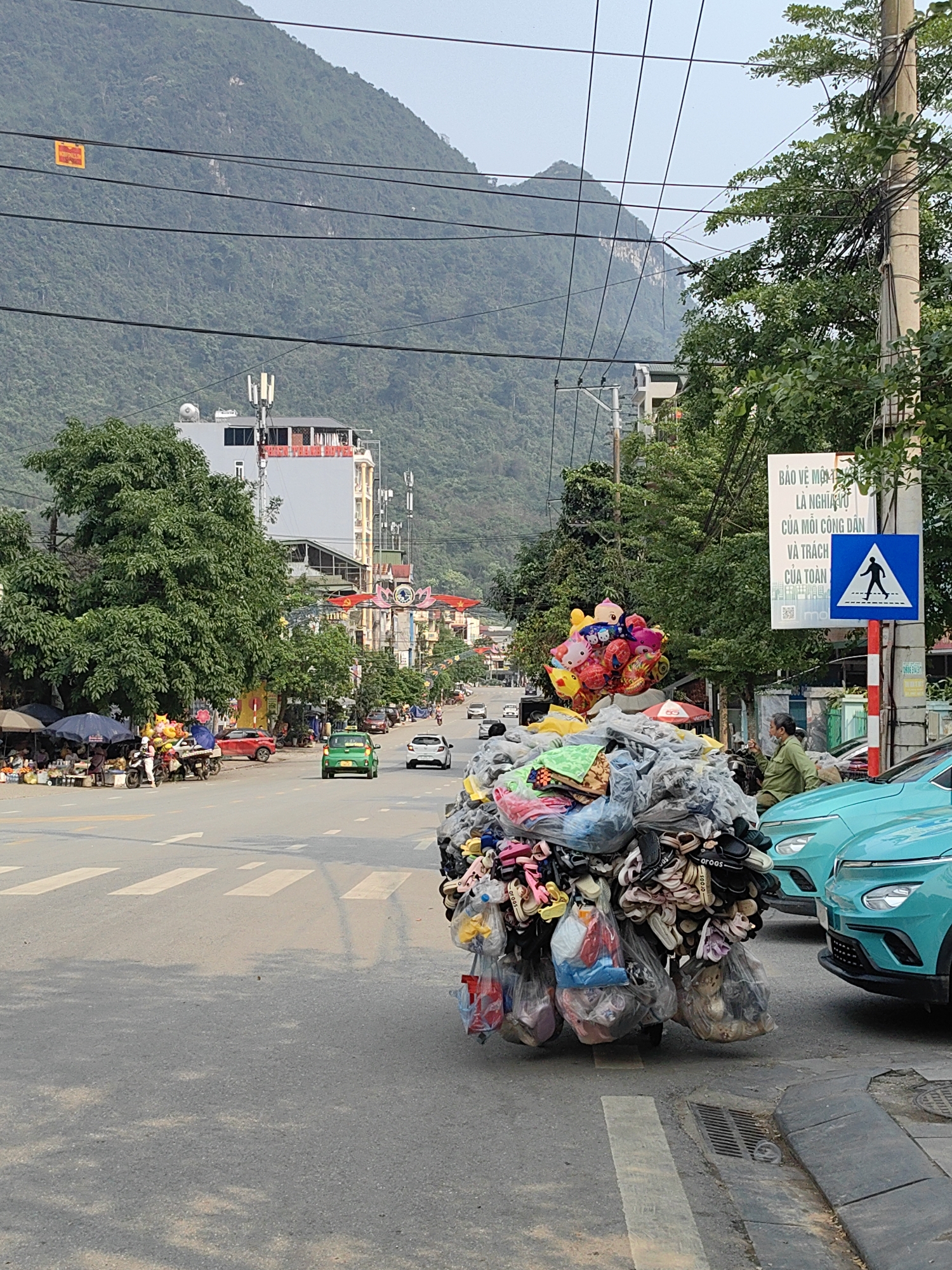 The image depicts a street scene in what appears to be Thành phố Hà Giang, Vietnam, with a street vendor prominently featured in the foreground. The vendor's cart is heavily laden with what appears to be shoes and other goods, topped with a bunch of colorful balloons. The cart is positioned near the edge of a crosswalk on a paved road. Two light blue cars are parked alongside the road to the right of the vendor's cart.
In the middle ground, traffic flows along the road, including a green car and a white car. A market area is visible on the left side of the road, with people gathered around stalls shaded by umbrellas.
In the background, a large mountain dominates the horizon, covered in lush green vegetation. Buildings and other infrastructure line the street further back, including a gray building with antenna arrays on its roof and a decorative archway across the street. Power lines are strung across the sky.
On the right side of the road, a sign written in Vietnamese is visible, mounted on a post with a pedestrian crossing symbol. A person sits nearby on a motorbike. The overall impression is of a bustling street scene in a city nestled amidst a mountainous landscape.