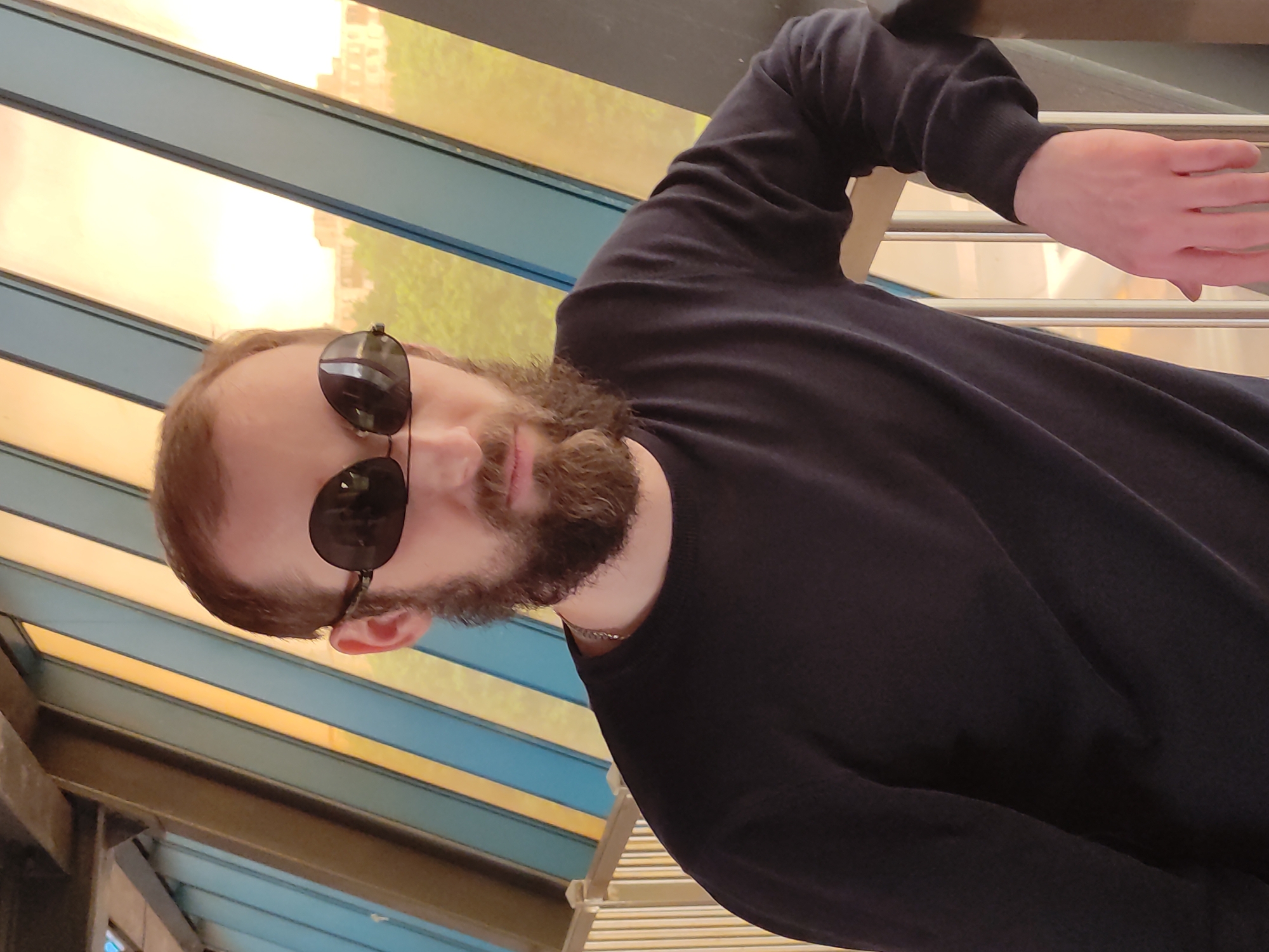 A man with a beard and sunglasses is photographed at an angle under a slatted roof with yellow and blue panels. He is wearing a black long-sleeved shirt. His right hand is resting on a metal railing. The location is likely in a city based on the presence of a building visible through the roof slats. Based on the style of the building visible through the roof and the overall environment, the city is likely Moscow, Russia.