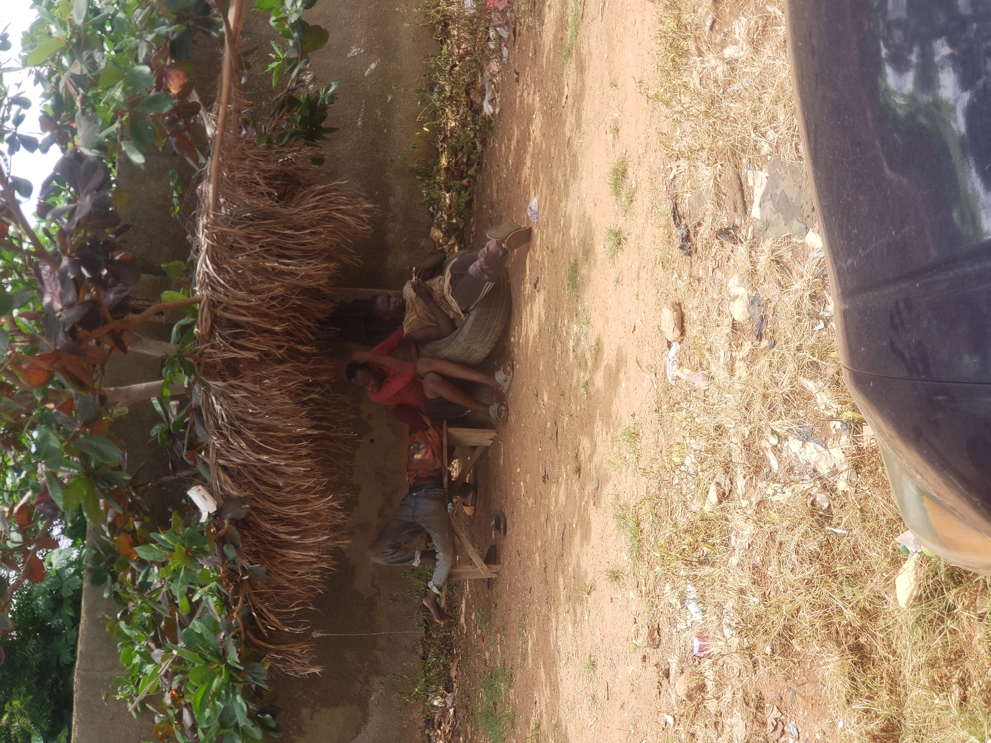 In Narayi, Nigeria, three individuals are resting under a makeshift shelter constructed from palm fronds and branches, providing shade from the sun. The shelter is situated against a textured, light-brown wall. One person lies horizontally on a wooden bench, with their head towards the left of the frame, their legs extended. Another person is seated upright on a tire to the right of the bench, looking down at a mobile device. A third person is seated on the bench next to the tire, leaning against the wall and appearing to be resting. The ground is composed of dry, reddish-brown soil with sparse patches of dry grass and scattered debris, including plastic bags and wrappers. In the foreground, the hood of a dark-colored vehicle is visible, partially obscuring the lower portion of the scene. Overhead, green foliage from a tree with reddish-brown leaves drapes over the shelter.