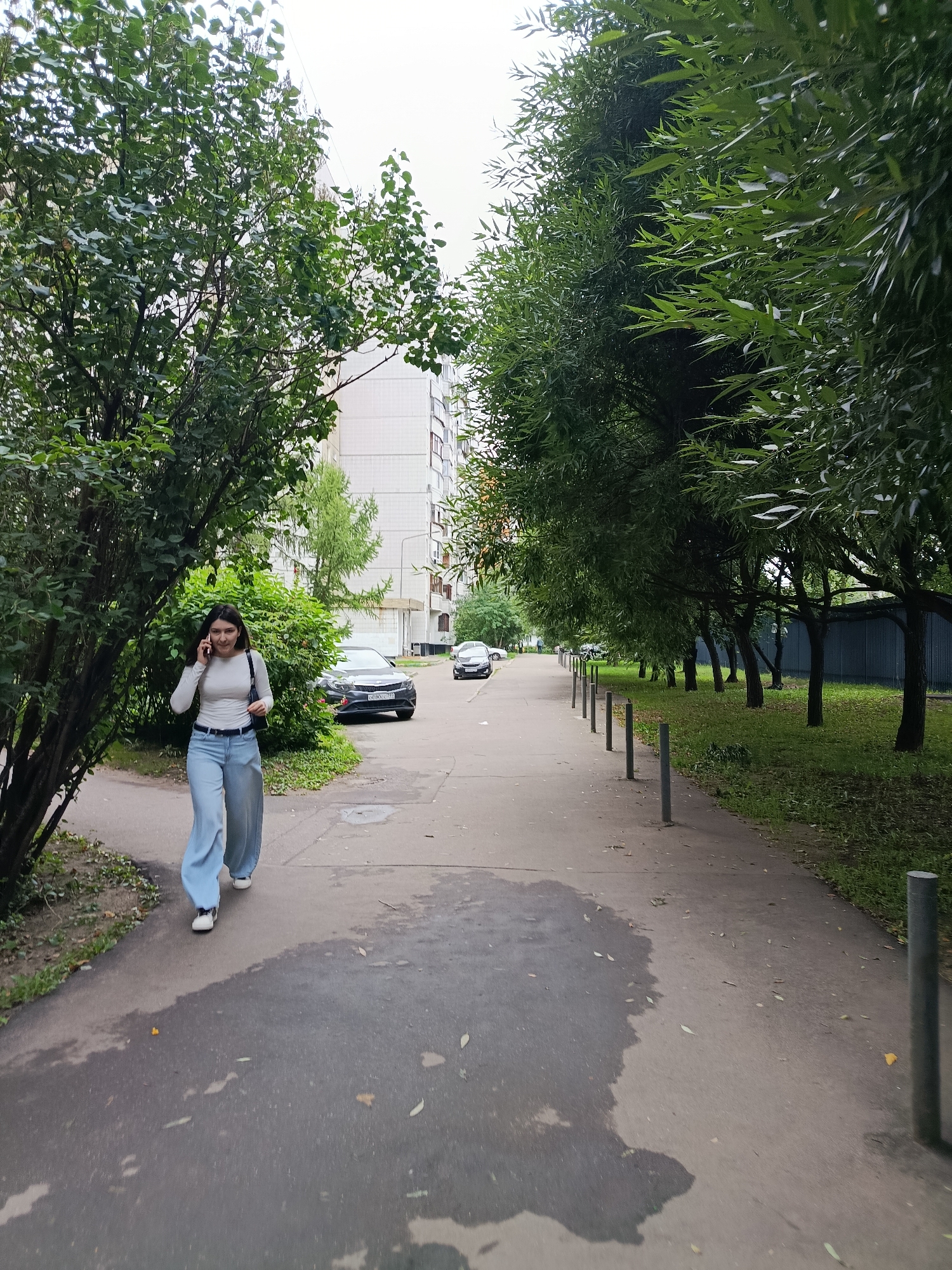 A young woman is walking on a sidewalk while talking on her phone in a residential area of Moscow, Russia. She is wearing a white long-sleeved shirt, light blue wide-leg jeans, and white sneakers. She has dark hair and is carrying a black bag on her left shoulder. The sidewalk is paved with concrete and has some wet patches. To her left, there are trees and a white apartment building. To her right, there is a row of trees and a metal fence. There are cars parked on the street in the background. The sky is overcast.