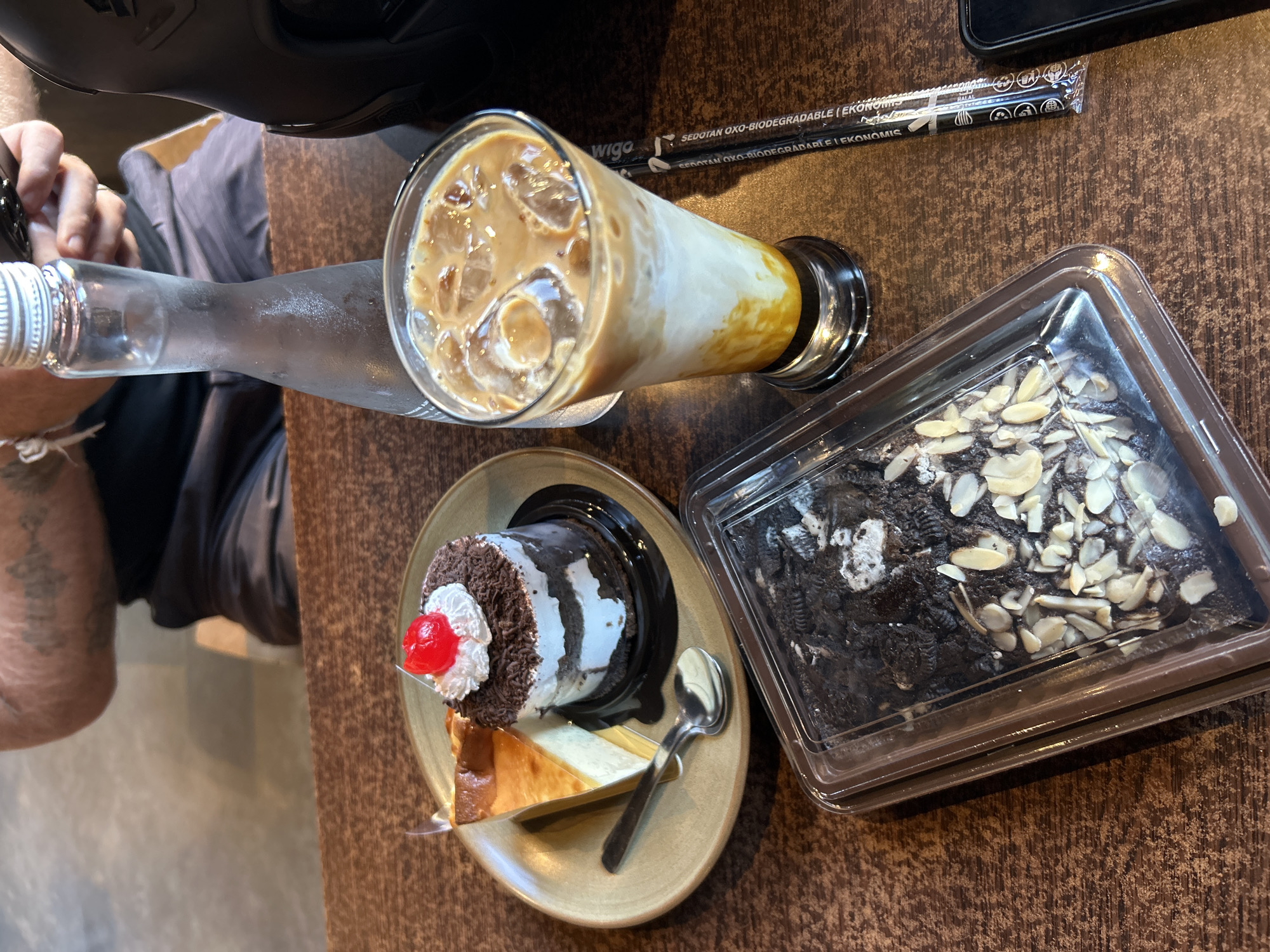 The image shows a close-up of a table with desserts and a drink, likely in a cafe or restaurant setting. On a brown plate, there is a slice of cheesecake and a small chocolate cake topped with whipped cream and a cherry. To the right of the plate, a tall glass contains an iced coffee drink with layers of milk and caramel sauce, topped with ice cubes. In the foreground, a rectangular clear plastic container holds a brownie topped with almonds and what appear to be crushed cookies. Next to the glass, a biodegradable straw is visible. A bottle of water stands behind the glass. In the background, a person's arm with tattoos is visible, suggesting they are sitting at the table. The table surface has a dark, wood-grain texture. The overall scene depicts a casual indulgence. The location is identified as Sukawati, Indonesia.