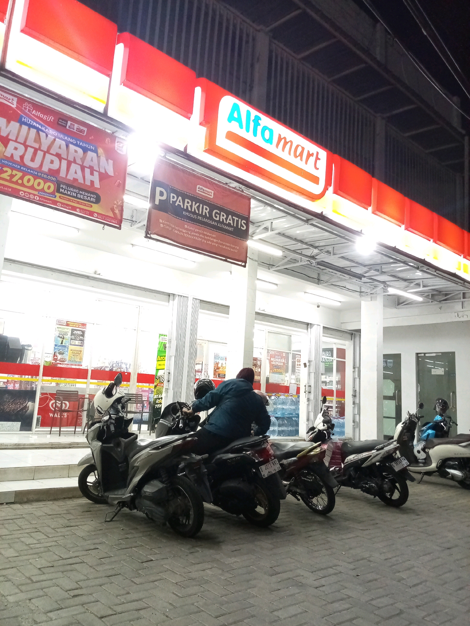 This is a street-level, medium-shot, eye-level photograph taken at night in front of an Alfamart convenience store. The store's facade is brightly lit by overhead fluorescent lights and the red and white Alfamart signage. In the foreground, several parked motorcycles are visible on a paved brick surface. One person, wearing a dark jacket, is seated on a scooter in the middle ground, facing away from the camera. Another person, also wearing a dark jacket and a helmet, is seated on a scooter to the right, facing the store entrance. The store entrance itself is glass-paned, allowing a view of shelves stocked with various products. Above the entrance, a banner advertises "PARKIR GRATIS" (Free Parking) for Alfamart customers. Another banner further to the left advertises "MILYARAN RUPIAH" (Billions of Rupiah) with a lottery promotion. The overall scene depicts a common urban nighttime setting outside a retail establishment in Bekasi, Indonesia.