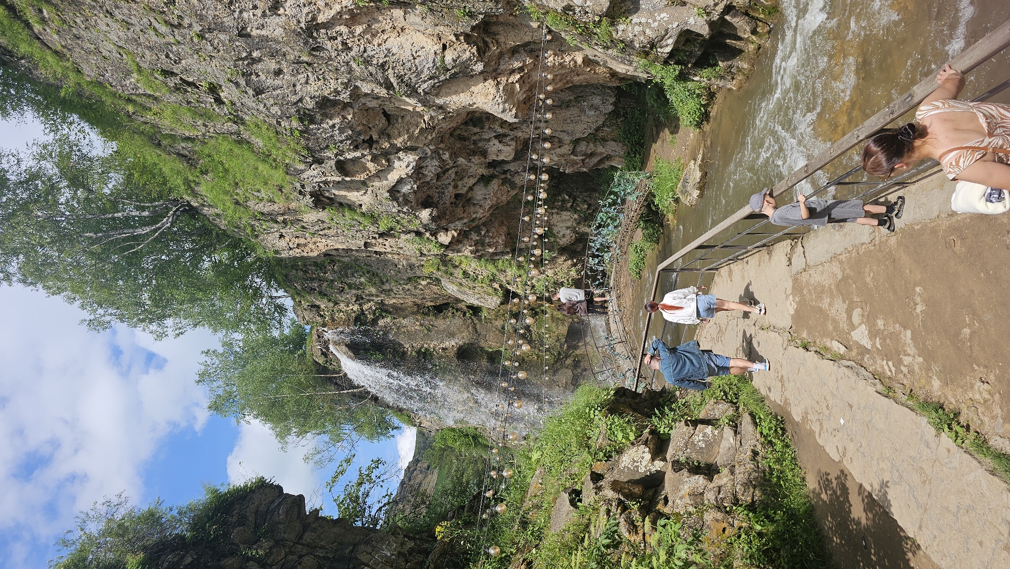 The image depicts a scenic natural landscape with a waterfall cascading down a rocky cliff face. A river flows through the scene, with a pedestrian bridge spanning across it. String lights are strung across the gorge, adding a decorative element. Several people are present, walking along a path and on the bridge. The environment is characterized by lush greenery and rugged rock formations, under a bright blue sky with scattered clouds. The location is identified as Красный Курган, Russia.