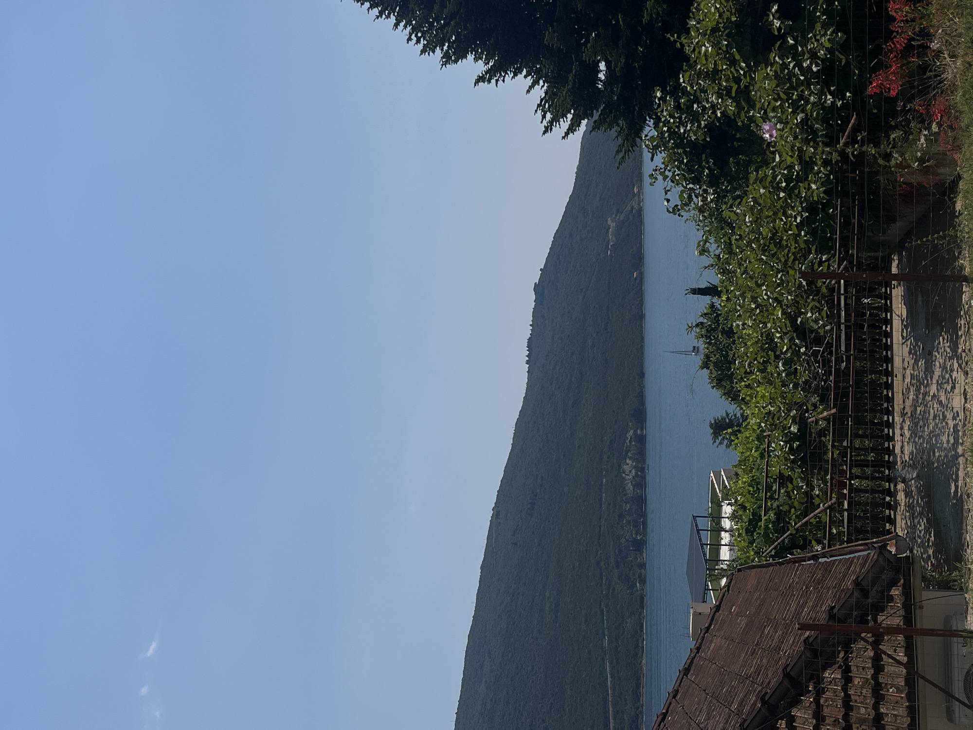 The image depicts a serene daytime scene in Kumbor, Montenegro, likely in late summer or early autumn given the lush greenery and clear sky. The vantage point is from a slightly elevated position, looking out over a body of water towards a steep, verdant mountain slope. The mountain is covered in dense foliage, giving it a rich, dark green appearance. The water is a calm, deep blue, reflecting the clear sky.
In the foreground, to the right of the frame, is a rustic scene with a weathered, brown tiled roof of a building. A metal fence runs along the edge of the property, partially obscuring some of the foliage. There are shadows cast on the ground, suggesting the sun is moderately high in the sky, indicating it is likely midday.
A single sailboat is visible on the water, adding a touch of activity to the tranquil landscape. The overall mood is peaceful and idyllic, characteristic of a coastal or lakeside retreat. The presence of trees and vegetation in the foreground frames the view, enhancing the sense of being in a natural environment. There is no visible text in the image.