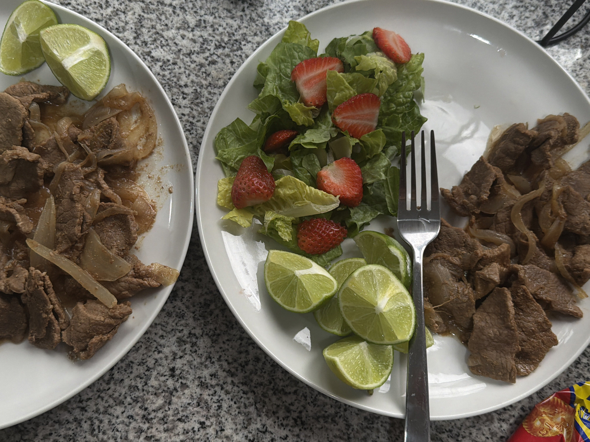 Two white plates are on a speckled gray countertop. The plate on the left contains sauteed beef and onions, with two lime wedges. The plate on the right also has sauteed beef and onions, along with a lettuce and strawberry salad, numerous lime wedges, and a silver fork. Part of a bag of chips is visible in the lower right corner. The location and city are unknown.