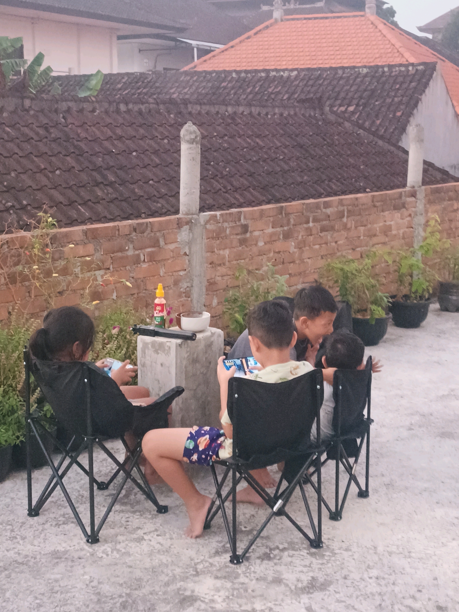 In Tabanan, Indonesia, four children are seated on black folding chairs on a concrete patio. Two children are facing each other, one looking at a mobile phone and the other engaged in conversation. Two other children are seated facing away from the camera, also looking at mobile phones. A small bottle of red liquid, a bowl, and a black object resembling a toy gun are placed on a concrete pedestal behind the children. The background features a brick wall, potted plants, and tiled rooftops.