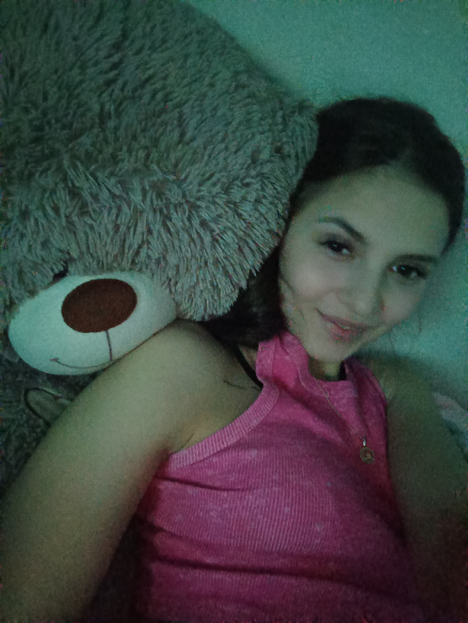 A young woman is depicted in a close-up, top-down shot, lying next to a large teddy bear. She is positioned on the right side of the frame, smiling softly and looking directly at the camera. She has dark hair and is wearing a pink ribbed tank top, along with a delicate necklace featuring a small, round pendant. Her bare shoulder and arm are visible, resting beside the teddy bear.
The teddy bear, a significant presence in the left and upper parts of the image, is large, fluffy, and appears to be grey or brownish-grey. Its face is partially visible, showing a white snout with a dark brown, circular nose and a subtle stitched smile. The overall scene suggests a cozy and relaxed moment, possibly in a bed or on a soft surface.
The setting is indoors, with an indistinct background that appears to be a wall or soft furnishing, characterized by a faint greenish hue. The lighting is subdued, contributing to a slightly grainy texture and a noticeable green/blue color cast across the entire image, which could be due to low light conditions or a photographic filter. The woman's expression conveys contentment. There is no visible text in the image.
Location: Unknown, Unknown