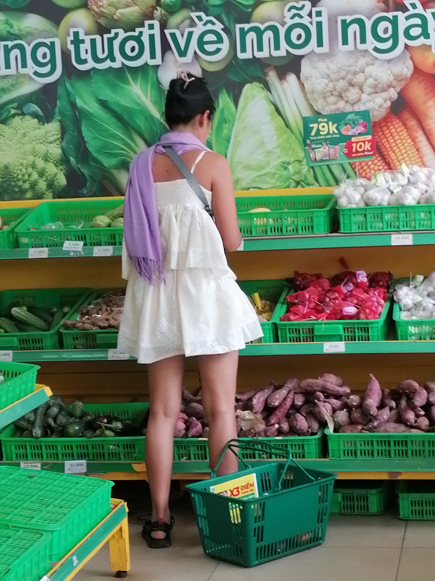 In a supermarket in Xuan Thanh, Vietnam, a young woman with dark hair and a light-colored dress, wearing a purple scarf, is browsing for produce. She is standing in an aisle with shelves stacked with various vegetables. The shelves are lined with green plastic crates containing items such as leafy greens, cucumbers, and onions. In front of her, a green shopping basket is placed on the floor. In the background, a large banner displays images of fresh vegetables and text in Vietnamese. The lighting appears to be typical of an indoor retail environment.