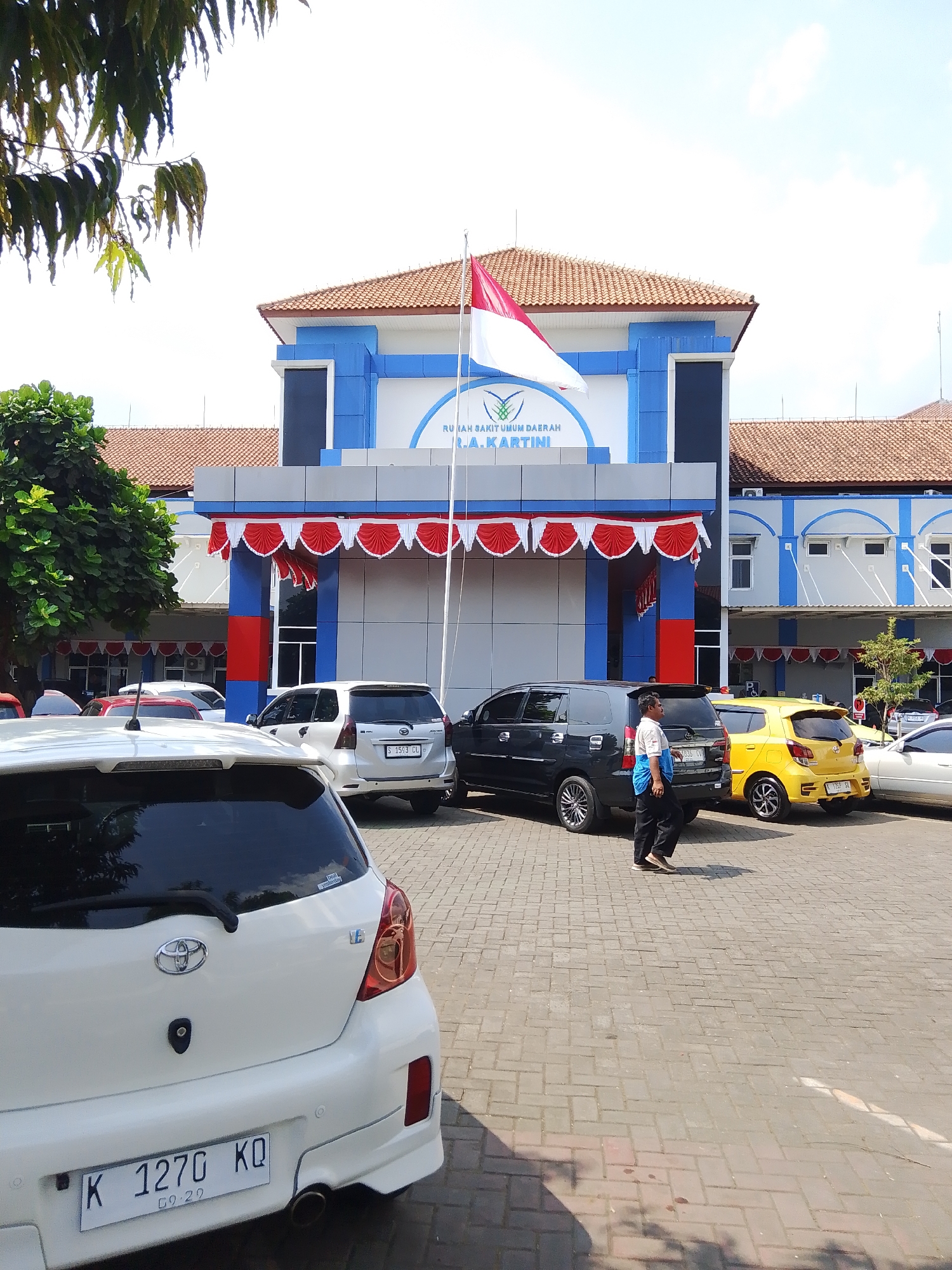 In Potroyudan, Indonesia, a building with blue and white accents, identified by the signage "RUMAH SAKIT UMUM DAERAH R.A. KARTINI," stands prominently. A red and white Indonesian flag flies from a flagpole in front of the building. The facade is decorated with a festive red and white draped banner. In the foreground, a white Toyota hatchback with license plate "K 1270 KQ" is parked on a paved surface. Adjacent to it are a black minivan and a yellow compact car. A man wearing a blue shirt and black pants walks across the parking lot. Trees with green foliage are visible to the left of the building, and the sky above is bright and clear.