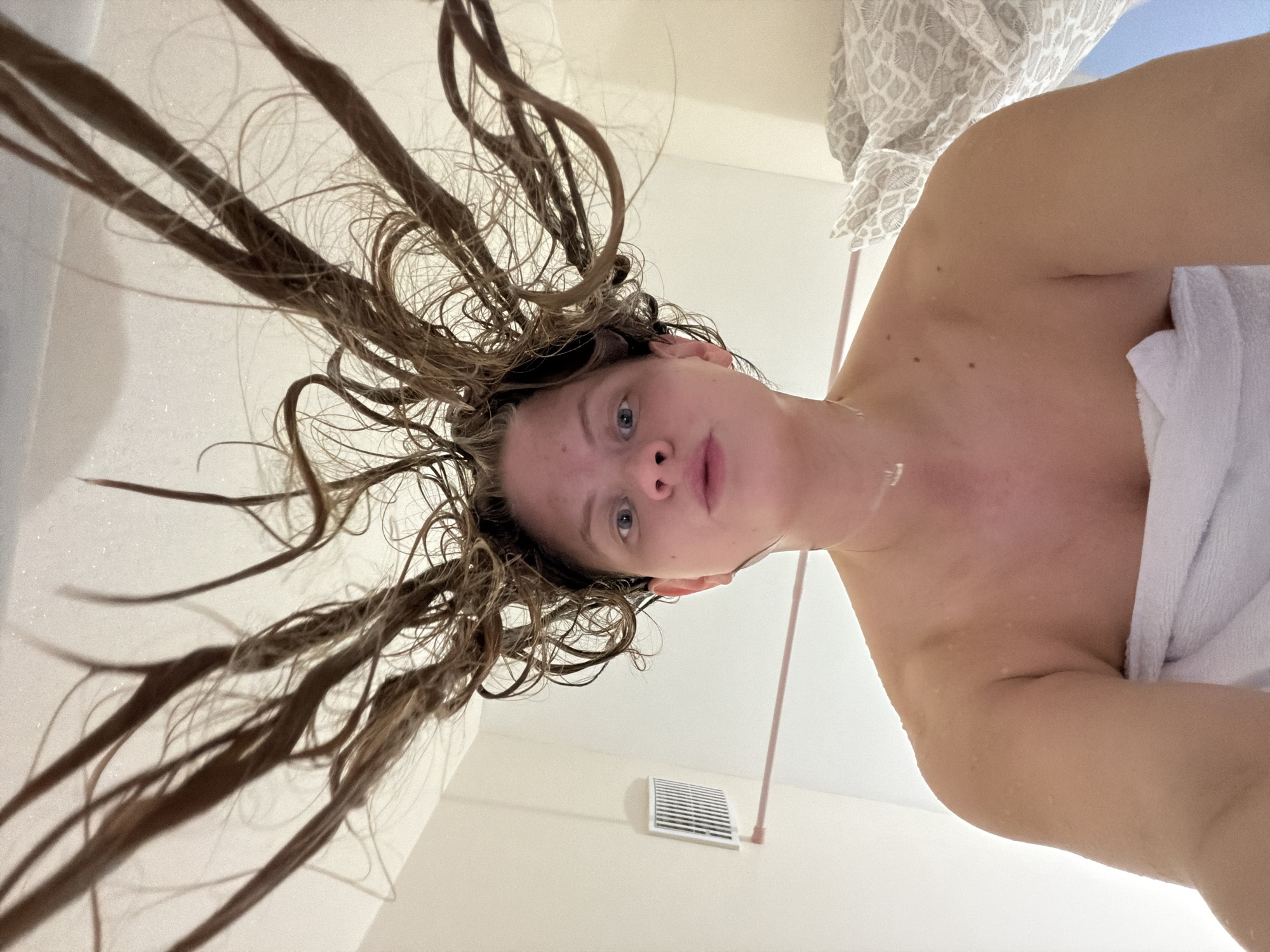This image, taken in Izhevsk, Russia, captures a person from a low-angle perspective, looking upwards directly at the camera. The subject is a young woman with fair skin and wet, light brown hair that appears to be fanned out and extending upwards, suggesting she is either inverted or leaning back after washing her hair. Water droplets are visible on her skin and hair. She has blue eyes and a neutral, slightly contemplative expression. Her torso is partially covered by a white towel.
The setting appears to be indoors, likely a bathroom, given the wet hair, towel, and the presence of a ventilation grille on the light-colored wall below her face. A thin, light-colored pipe runs vertically on the wall near the grille. In the upper right corner, a patterned curtain or fabric is partially visible. No text is discernible in the image. The scene conveys a moment immediately following a shower or hair wash, presented with an unusual and dynamic perspective.