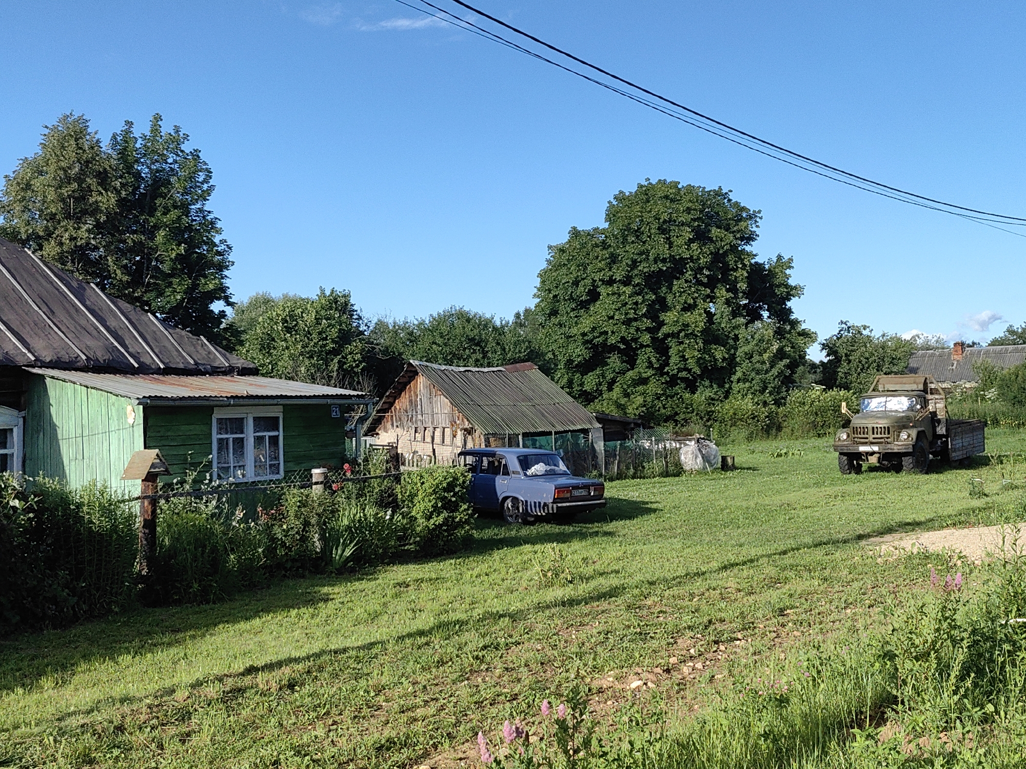The image shows a rural scene in the city of Квант-88 Снт, Russia. In the foreground, a lush green lawn slopes gently downwards, with patches of dry earth and scattered wildflowers. The middle ground features a green wooden house with a metal roof, a fence, and a bird feeder on a wooden post. Next to the house is a wooden shed with a corrugated metal roof. A blue sedan is parked in the yard. In the background, a large green military truck is parked on a dirt road. Tall trees and dense foliage surround the property, and a clear blue sky with a few wispy clouds forms the backdrop. There are no people visible in the image.