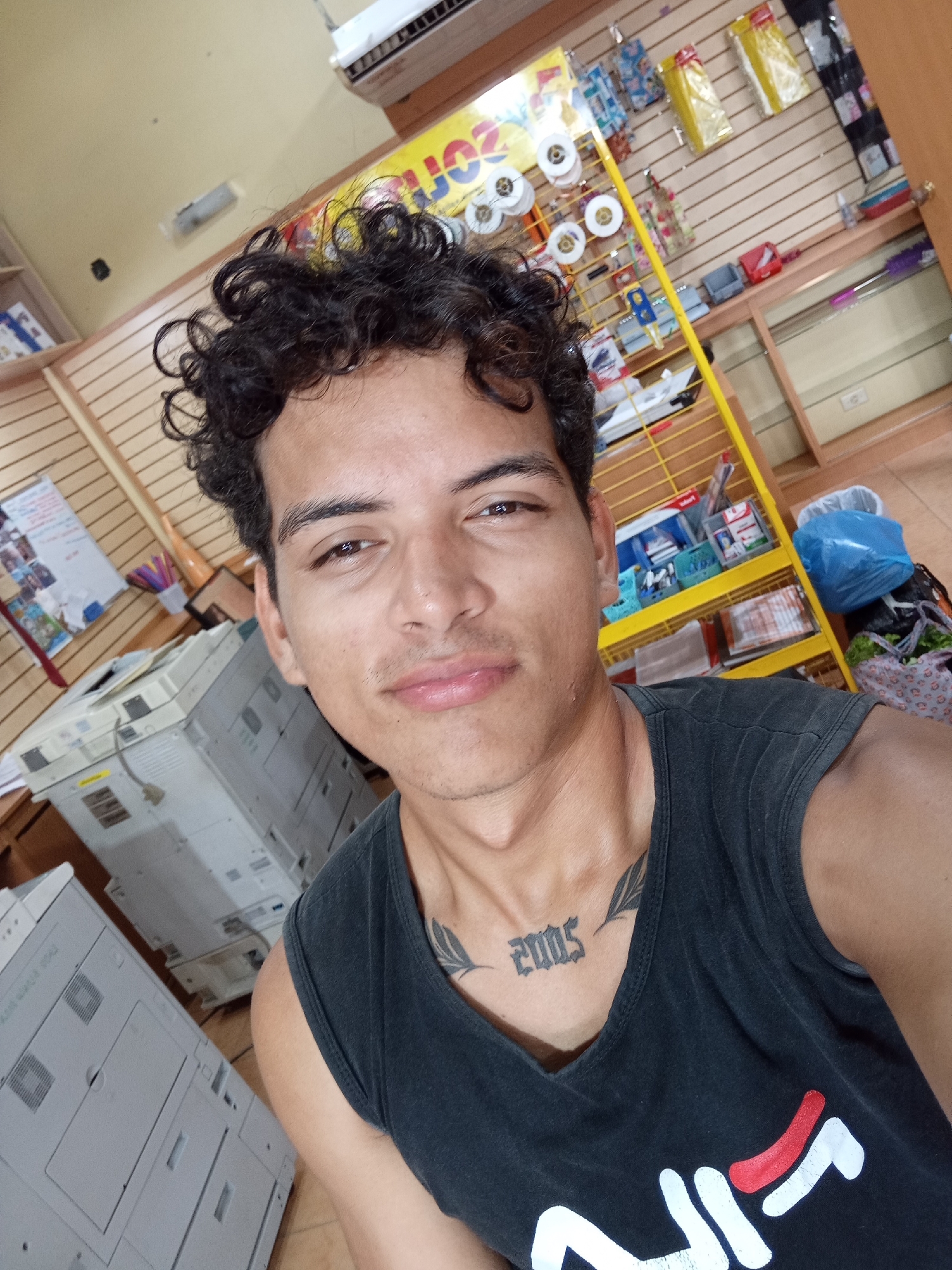 A young adult male is depicted in a close-up, chest-up selfie, positioned slightly off-center. He has dark, curly hair, dark eyes, and light stubble. He is wearing a black sleeveless top, which displays a portion of a white and red Fila logo on the lower front. Prominently visible on his upper chest is a tattoo of the year "2005" in a gothic script, accompanied by a partial feather or leaf design extending towards his left shoulder. His head is tilted slightly, and he has a subtle smile.
The background suggests an indoor commercial or office environment, likely a print shop or stationery store in Juan Griego, Venezuela. To the viewer's right, a bright yellow metal shelving unit with multiple tiers holds various retail items, including white spools of ribbon and plastic-wrapped packages. Behind this, a light brown wooden slatwall display system is visible. Further right, a section of empty wooden shelving and a power outlet are present, with a blue plastic bag and another patterned bag resting on the floor or a low shelf.
To the viewer's left, two large, light-grey office copier machines are positioned, indicating a printing service area. A wooden slatwall is also present on this side, featuring a white board with attached papers and a container of writing instruments. High on the wall in the upper left, an air conditioning unit is mounted. The ceiling is a light cream color. The overall lighting appears to be from overhead artificial sources.