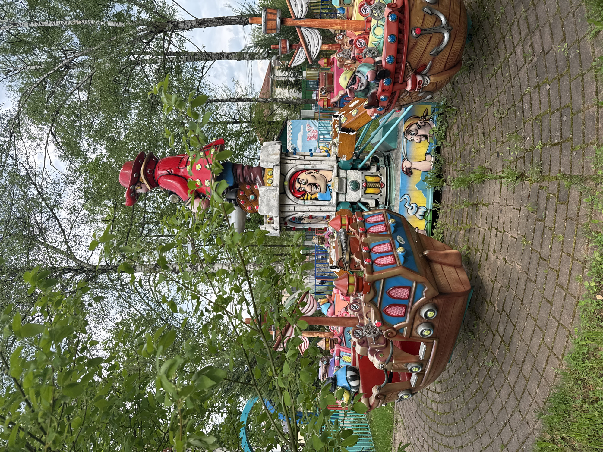 The image shows a carousel ride in Dmitrov, Russia, featuring pirate-themed boats and characters. The ride is located outdoors, with trees surrounding the area and a brick-paved path running alongside it. There is no visible people in the image.