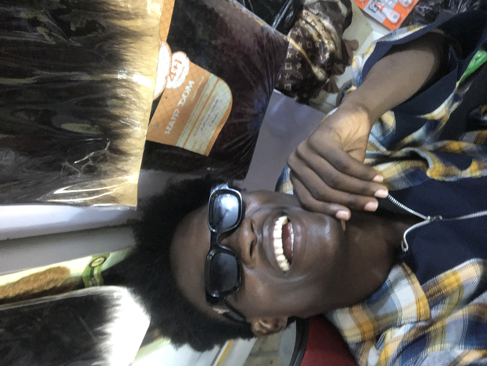 A dark-skinned individual is depicted in a close-up, chest-up vertical photograph taken within what appears to be a retail establishment in Ado, Nigeria. The individual possesses short, dark, textured hair and is wearing dark, rectangular sunglasses. A wide, open-mouthed smile is present, revealing both upper and lower teeth. The right hand is positioned with fingers near the chin. The individual is dressed in a plaid shirt featuring blue, yellow, and white patterns, worn over a dark blue garment with a zippered front. The background exhibits various commercial items. To the upper right, multiple large, transparent plastic bags containing dark hair extensions are visible, with one bag displaying a label that reads "HAIR DOM" and "4+1 Closure" in orange and white text, along with smaller text indicating hair types such as "SILKY STRAIGHT" and "JERRY CURL." To the upper left and above the individual's head, obscured items, possibly rolls of material or additional hair products, include segments of white and golden-hued objects. In the lower right background, a brown fabric patterned with lighter motifs, resembling a designer print, is present, alongside a clear plastic package containing several small, white, circular objects.