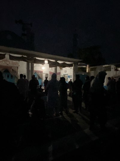 The image shows a group of people gathered outside a building at night. The building appears to be a mosque based on the dome and minaret-like structure visible in the upper part of the frame. Individuals stand in the dim light emanating from within the building. The location is possibly Kandahar, Afghanistan, given the cultural context of the building and the clothing of the people present.