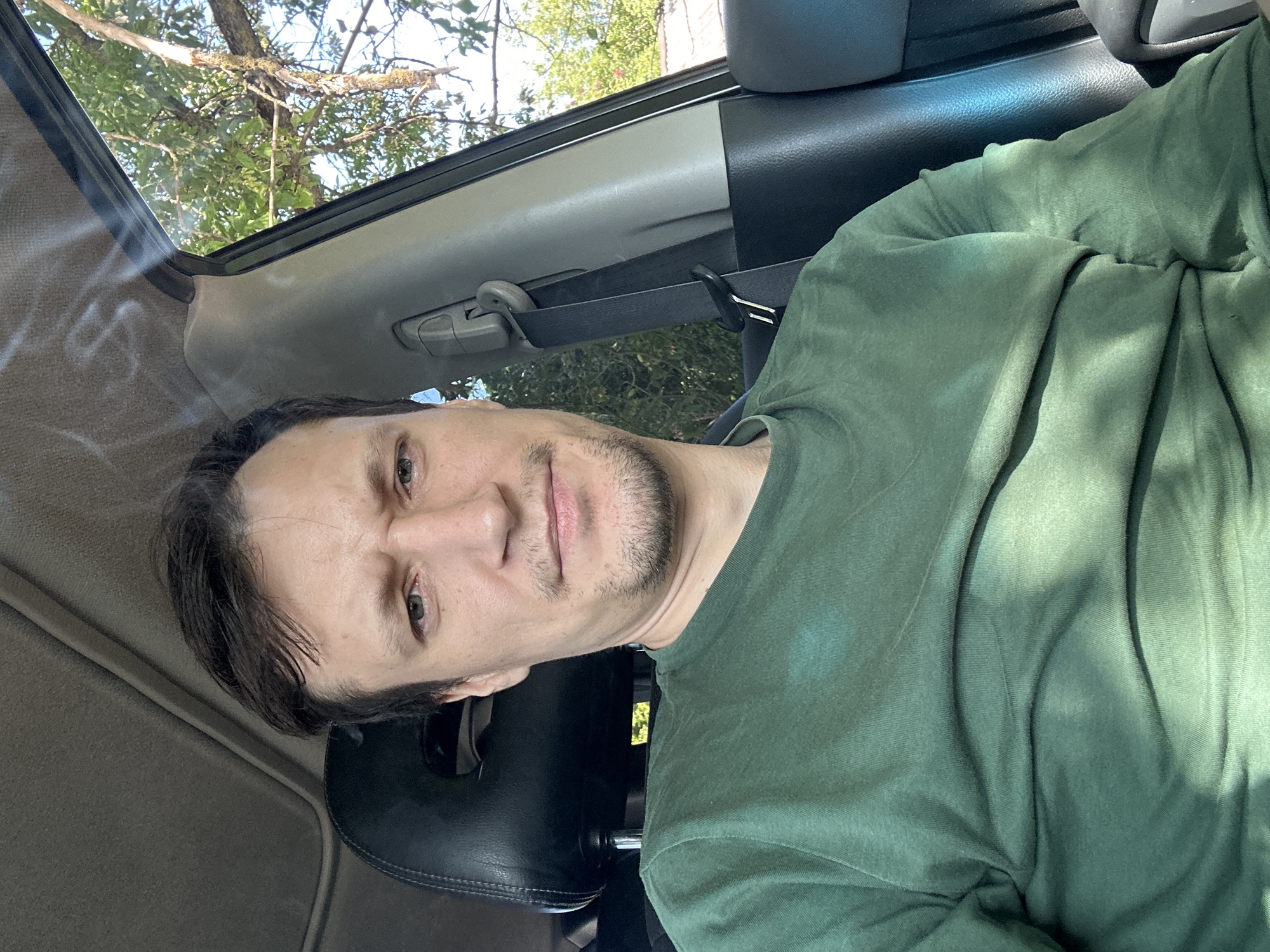 A man is captured in a selfie from the chest up, seated in the driver's seat of a vehicle. He is wearing a green long-sleeved shirt. Sunlight filters through the car's windows, casting shadows across his face and clothing. The interior of the car is visible, with a dark leather seat, seatbelt, and part of the dashboard. Through the open window, trees and a clear sky suggest an outdoor setting, possibly in Makiv, Ukraine, given the context. The man's expression is neutral, with a slight frown. There is a faint plume of smoke visible in the upper portion of the image, suggesting the presence of a cigarette or similar object.