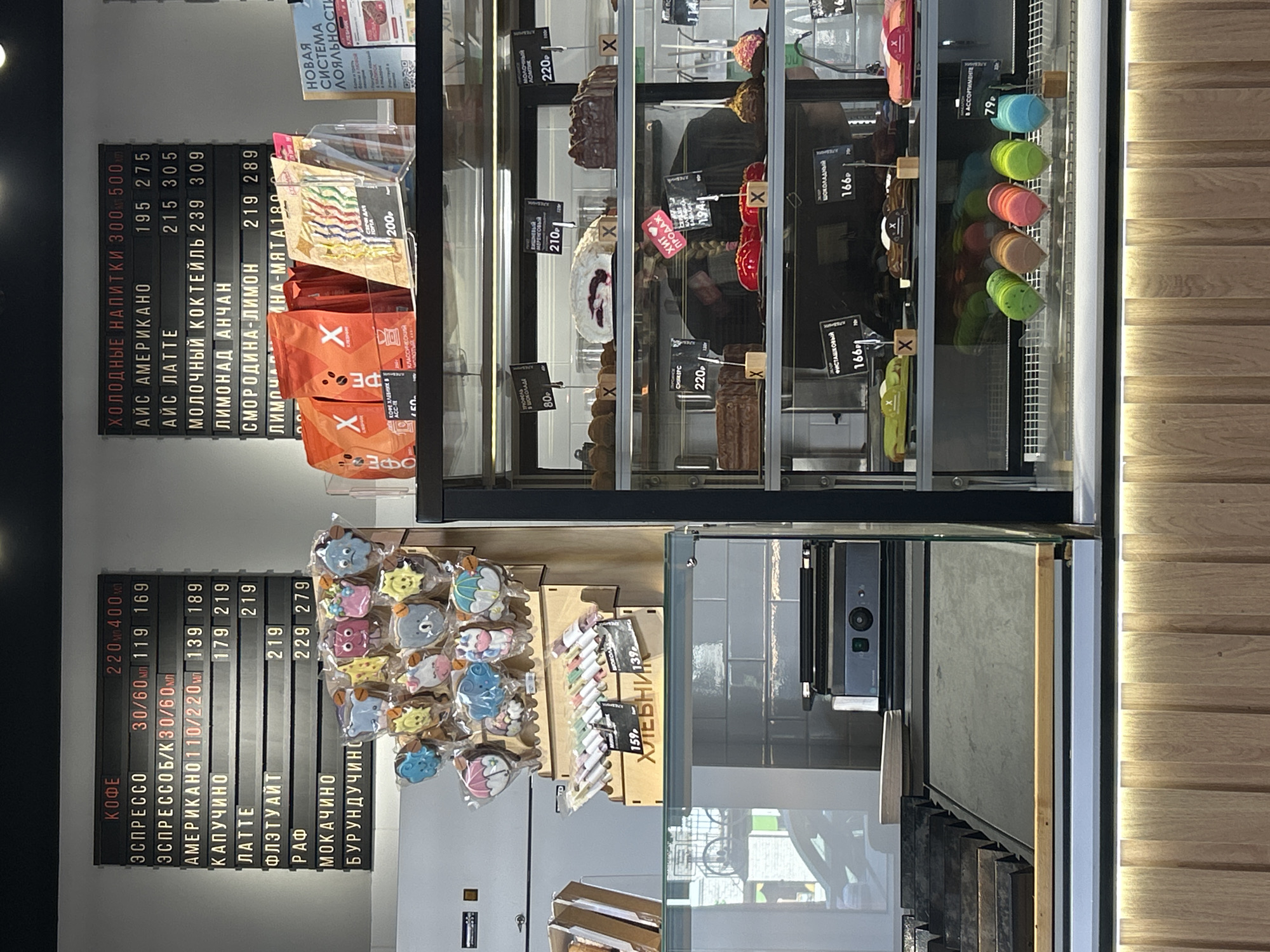 The image shows the interior of a cafe in Yanino-1, Russia, specifically a display case filled with various pastries and a menu board. The menu board on the left lists coffee drinks with prices for different sizes. The menu board on the right lists cold drinks with prices. The display case contains rows of colorful macarons, eclairs, cakes, and other sweets. Some items are individually priced, with labels such as "шоколадный" (chocolate), "фисташковый" (pistachio), and "ассортименте" (assortment). There are also bags of coffee beans and packaged cookies on display. The overall environment appears to be a clean and modern cafe setting. No people are visible in the image.