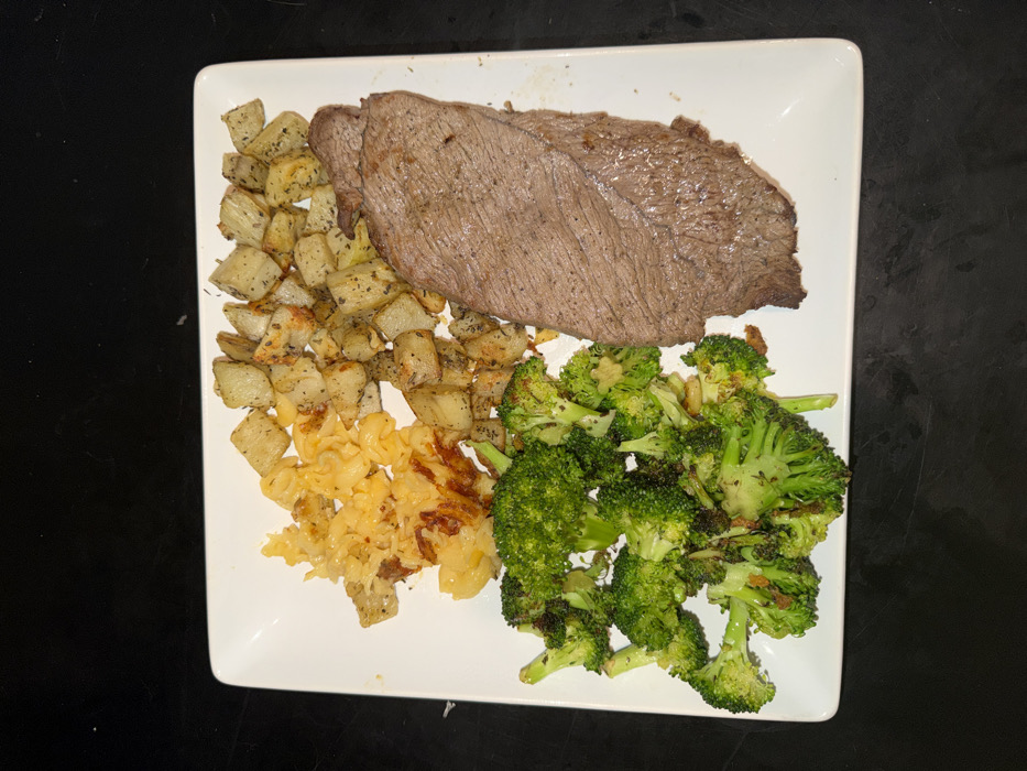 A white rectangular plate, with slightly rounded corners, holds a complete meal and is centered on a dark, uniform surface. The plate contains four distinct food items. On the right, a large, un-sliced piece of cooked beef steak, brown-grey in color, rests. Above it, in the top-center, is a serving of cubed, roasted potatoes, golden-brown and seasoned with visible herbs. To the left of the potatoes, in the top-left quadrant, is a portion of macaroni and cheese, consisting of elbow pasta coated in melted, golden-orange cheese, with some sections exhibiting a crisp, browned texture. In the bottom-left, a quantity of roasted green broccoli florets, some with slightly browned edges, completes the meal. No individuals, actions, or interactions are depicted. The scene is located in Los Angeles, United States of America.