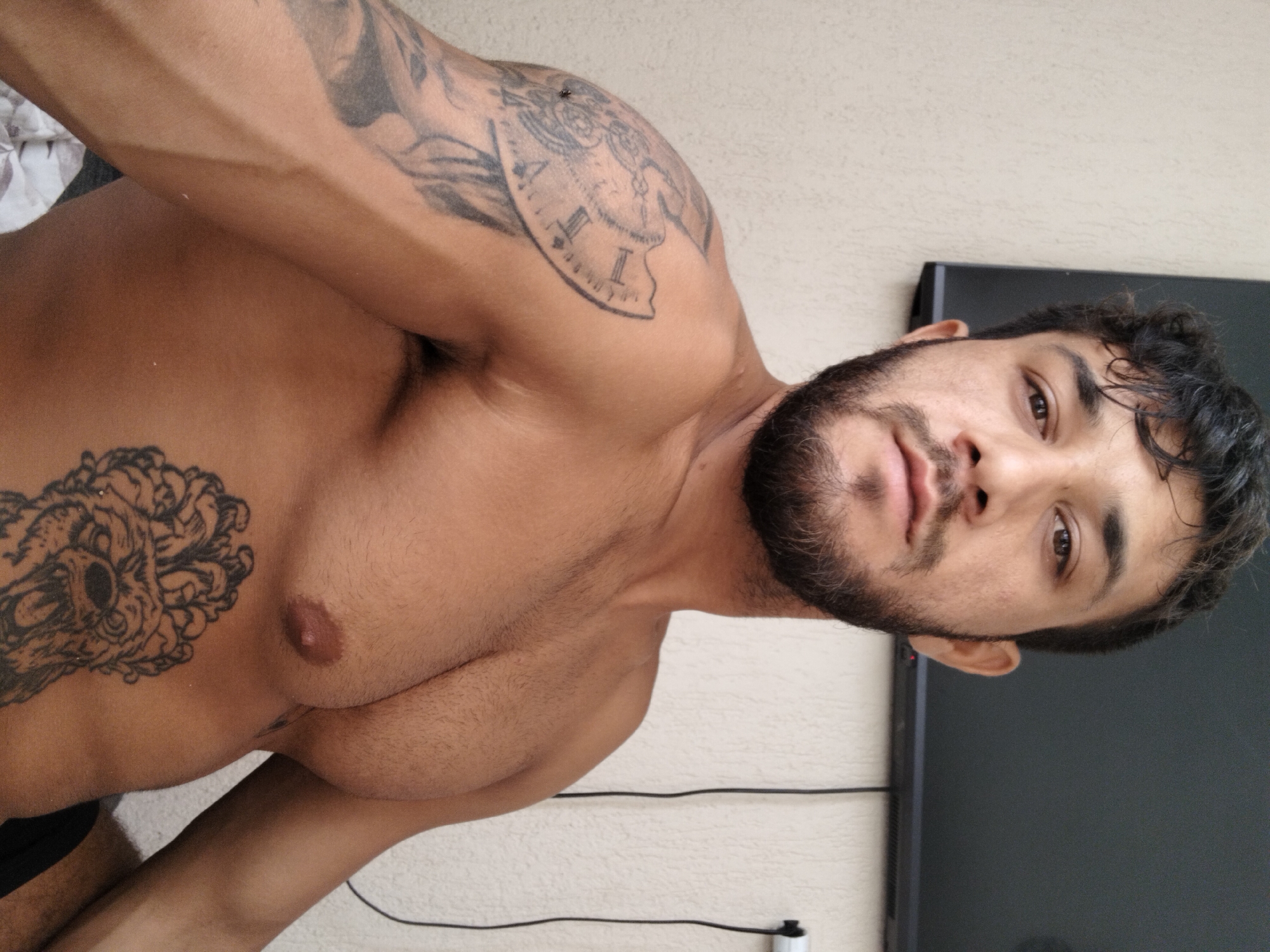 A man with dark curly hair and a beard, shirtless, is posing for a photo. He has tattoos on his left shoulder and chest, depicting a clock face and a lion. The background appears to be an indoor setting with a wall and a television screen mounted above him. The lighting is natural, coming from the front. The man is looking directly at the camera. The scene is not located in Tsarevo, Bulgaria.