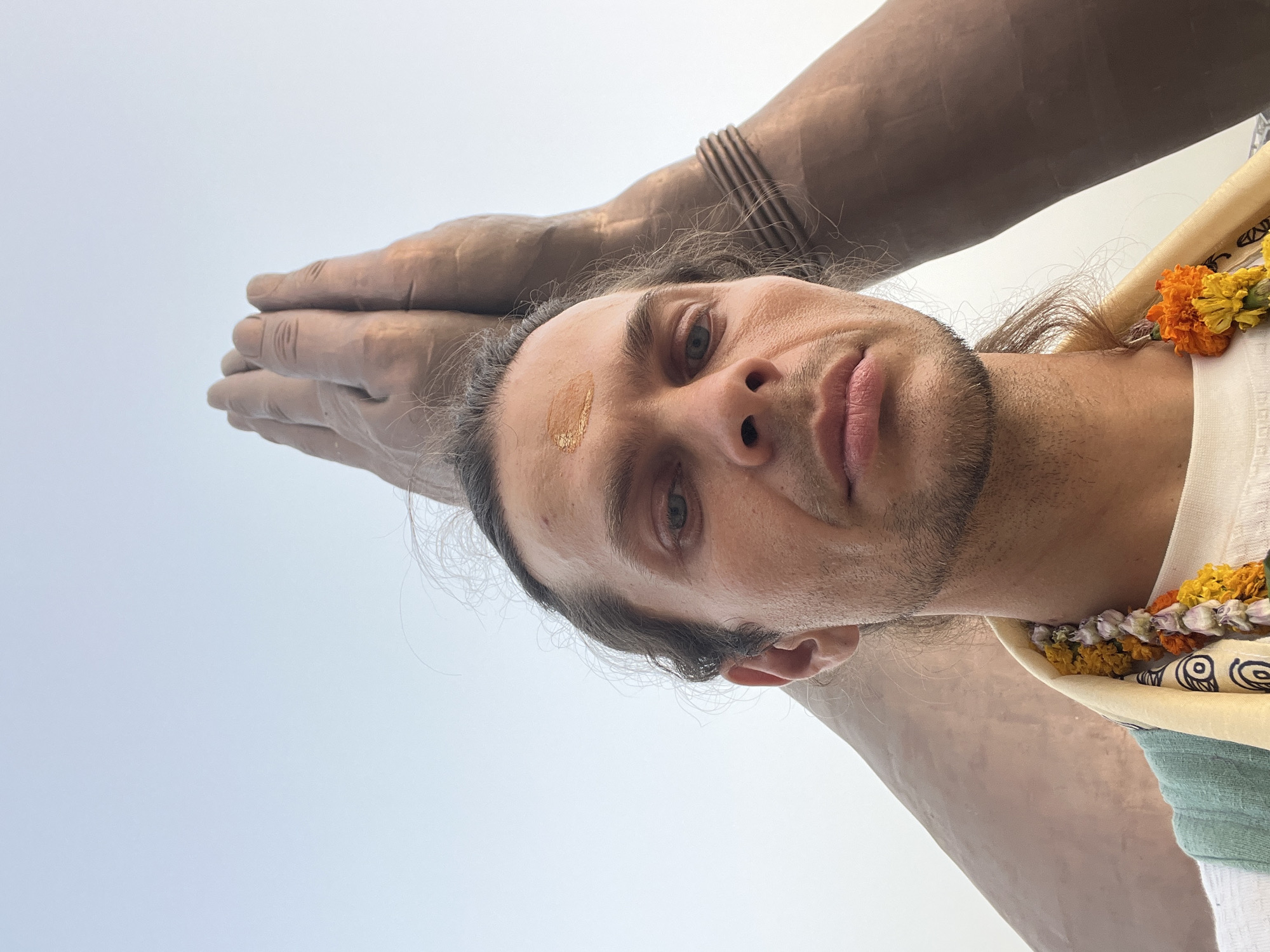 A medium shot shows a fair-skinned person with shoulder-length brown hair, a beard, and stubble under his lip. They are wearing a white garment with a patterned yellow scarf draped over their left shoulder adorned with orange and white flowers. The person has a yellow-gold dot on their forehead. Hovering over the person's head are two clasped bronze-colored hands. Behind the person is a pale, light blue sky. This image was taken in Mughalsarai, India.