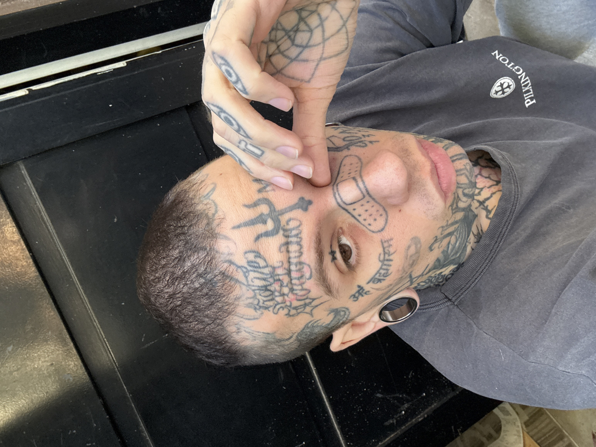 A person, likely male, is captured in a close-up, slightly tilted portrait, possibly lying down or leaning back. The individual has extensive black ink tattoos covering their face, hands, and neck. Their hair is short and dark, and a large silver ear gauge is visible in their left earlobe.
The person is wearing a grey, possibly faded, long-sleeved sweatshirt. On the right side of the chest (viewer's left), the word "PILKINGTON" is embroidered vertically, accompanied by a circular logo featuring a stylized cross or shield design.
Notable facial tattoos include a trishul (trident) symbol on the forehead, a "bandage" design on the nose, and various script tattoos. On the person's left cheek (viewer's right), a prominent tattoo reads "GAY". On the right cheek (viewer's left), the Sanskrit phrase "अहं ब्रह्मास्मि" (Aham Brahmasmi), meaning "I am Brahman" or "I am the Absolute Reality," is visible. The person's right hand, also heavily tattooed with geometric patterns and script, is gently touching their temple near the left eye.
The person looks directly at the camera with a neutral to contemplative expression. The background consists of a dark, possibly black, surface, which could be a table or floor, with some specks of dust or debris. The lighting appears to be soft and even, suggesting an indoor setting. No specific activity beyond posing for a photograph is discernible. The scene is set in Buenos Aires, Argentina.