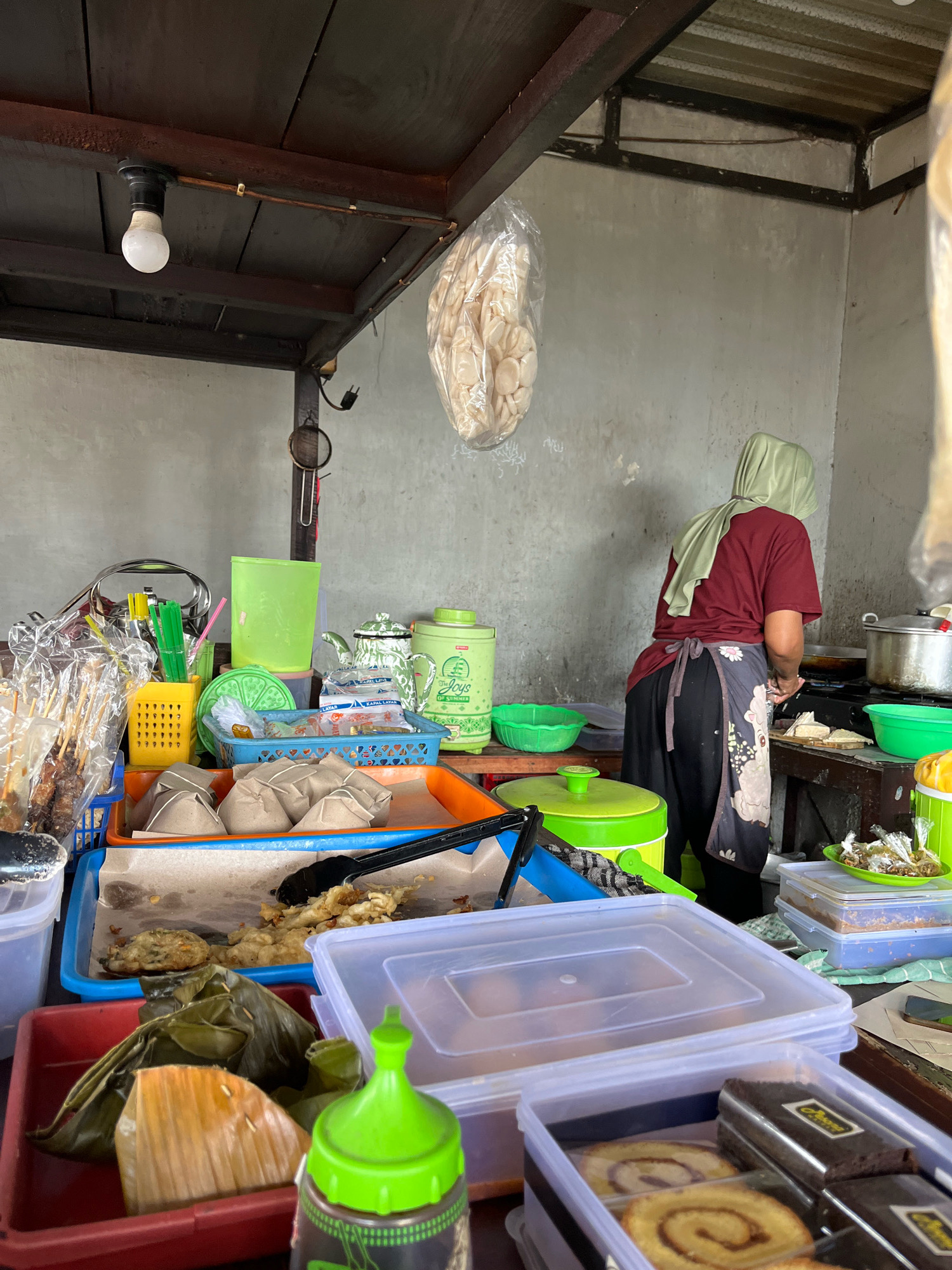 A food stall in Banguntapan, Indonesia, is depicted. A woman wearing a maroon shirt, a dark apron with a floral design, and a light green hijab is standing and cooking. She is facing away from the camera and appears to be preparing food on a stovetop, with pots and pans visible. The stall is filled with various food items and containers. In the foreground, there are plastic containers holding rolled cakes and brownies, and a red container with food wrapped in banana leaves. To the left, a blue tray holds wrapped food parcels and some fried items, accompanied by tongs. Other items on display include skewers of food in plastic packaging, a yellow utensil holder with green straws, a blue basket with more wrapped food, a green pitcher, and a green thermos. A light bulb hangs from the wooden ceiling. The background consists of a concrete wall.