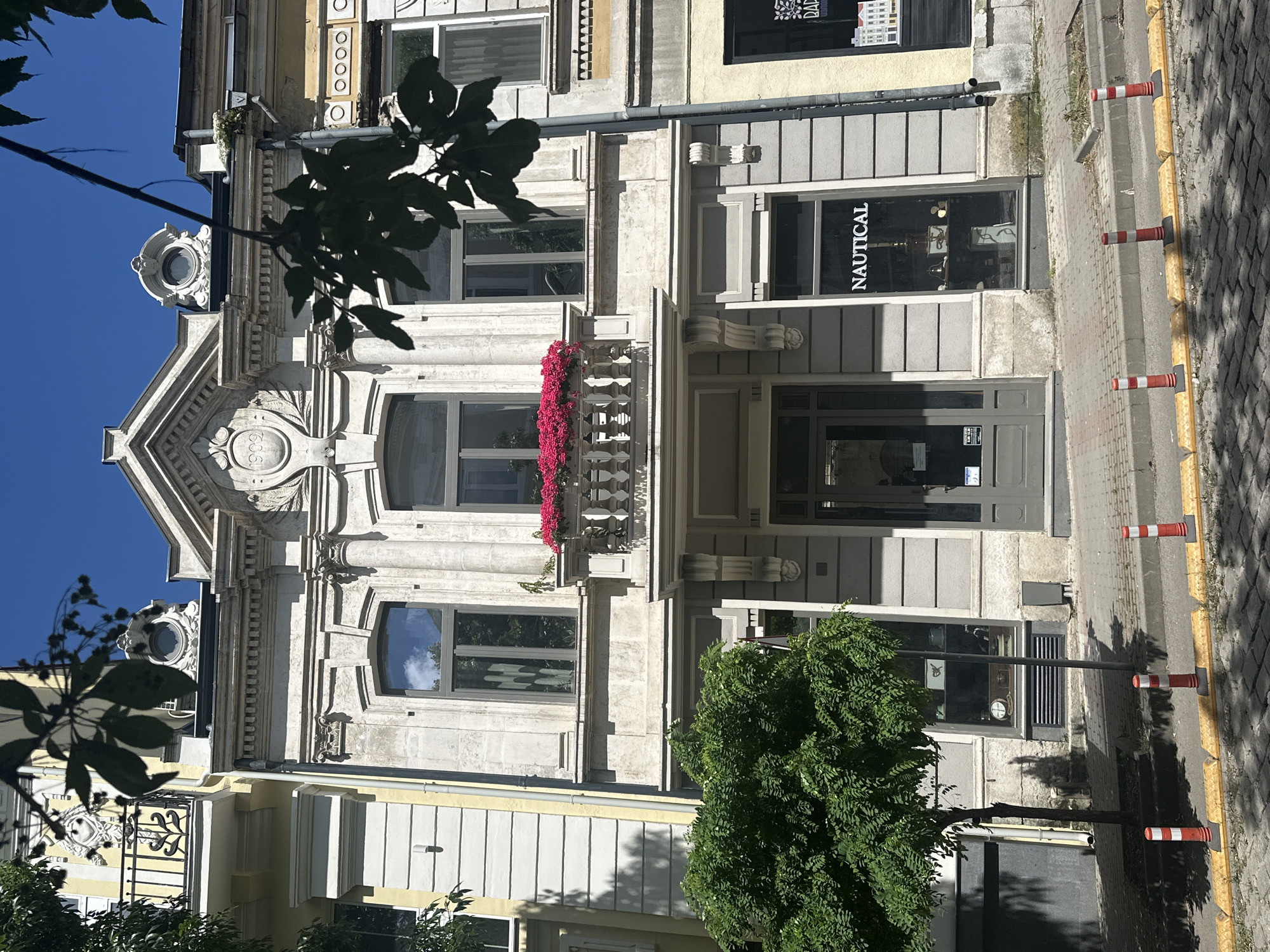 The image shows the exterior of a building on a street in an unknown city. The building has a light-colored facade with classical architectural details, including columns, decorative carvings, and a balcony with pink flowers. The ground floor features storefronts. One storefront has a sign that reads "NAUTICAL." The street is paved with cobblestones and there are several traffic bollards visible. A green tree is planted in front of the building. The sky is blue and clear.
