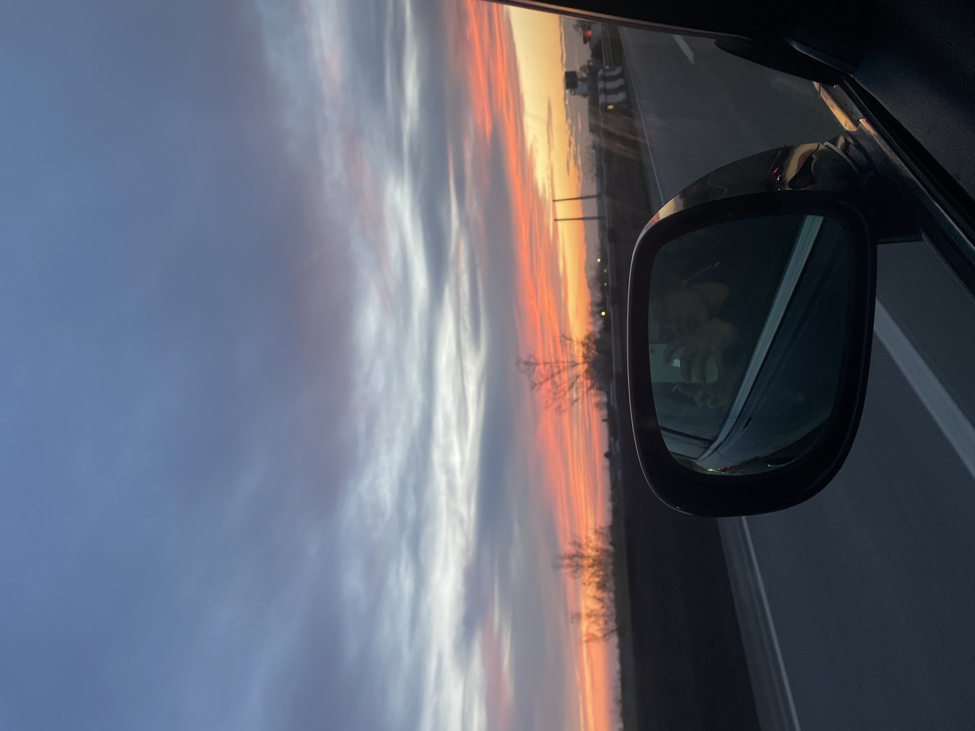 The image, likely taken from inside a moving car, features a side mirror reflecting a sunset view. The sky displays a mix of colors, transitioning from a deep blue to orange and hints of red near the horizon. Clouds are present, streaking across the sky. Bare trees or branches are visible along the horizon line, adding a silhouette effect. The mirror's reflection also shows part of a road with lane markings. The car interior, including the door frame and window, is visible around the mirror.
The location is likely in the area of Bolzano / Bolzan, Italy, given the request. This inference is based solely on the prompt's instruction, as there are no specific geographical markers in the image itself.