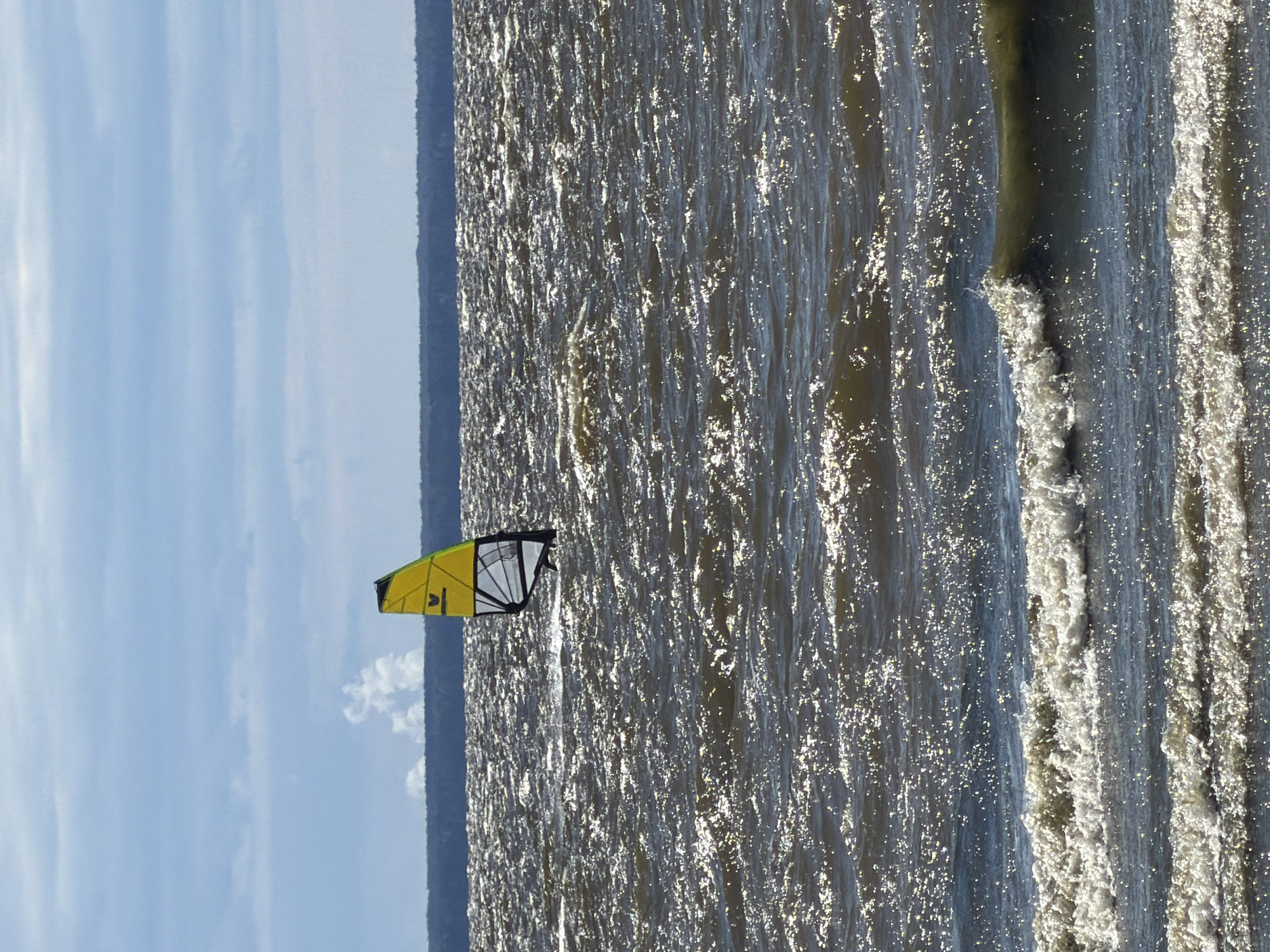 A solitary windsurfer is engaged in their sport on a choppy body of water, likely the Gulf of Finland, near Saint Petersburg, Russia. The windsurfer is positioned in the middle distance, on the right side of the frame, with their yellow and white sail prominently displayed against a backdrop of a dark, forested horizon and a hazy blue sky with scattered clouds. The water's surface is heavily textured with numerous small waves and glinting sunlight, suggesting a breezy day. In the far distance, on the horizon, a faint plume of smoke or mist rises from the land, possibly indicating industrial activity or natural atmospheric conditions. The foreground of the image features larger, white-capped waves rolling towards the viewer, adding to the sense of motion and dynamism of the scene.