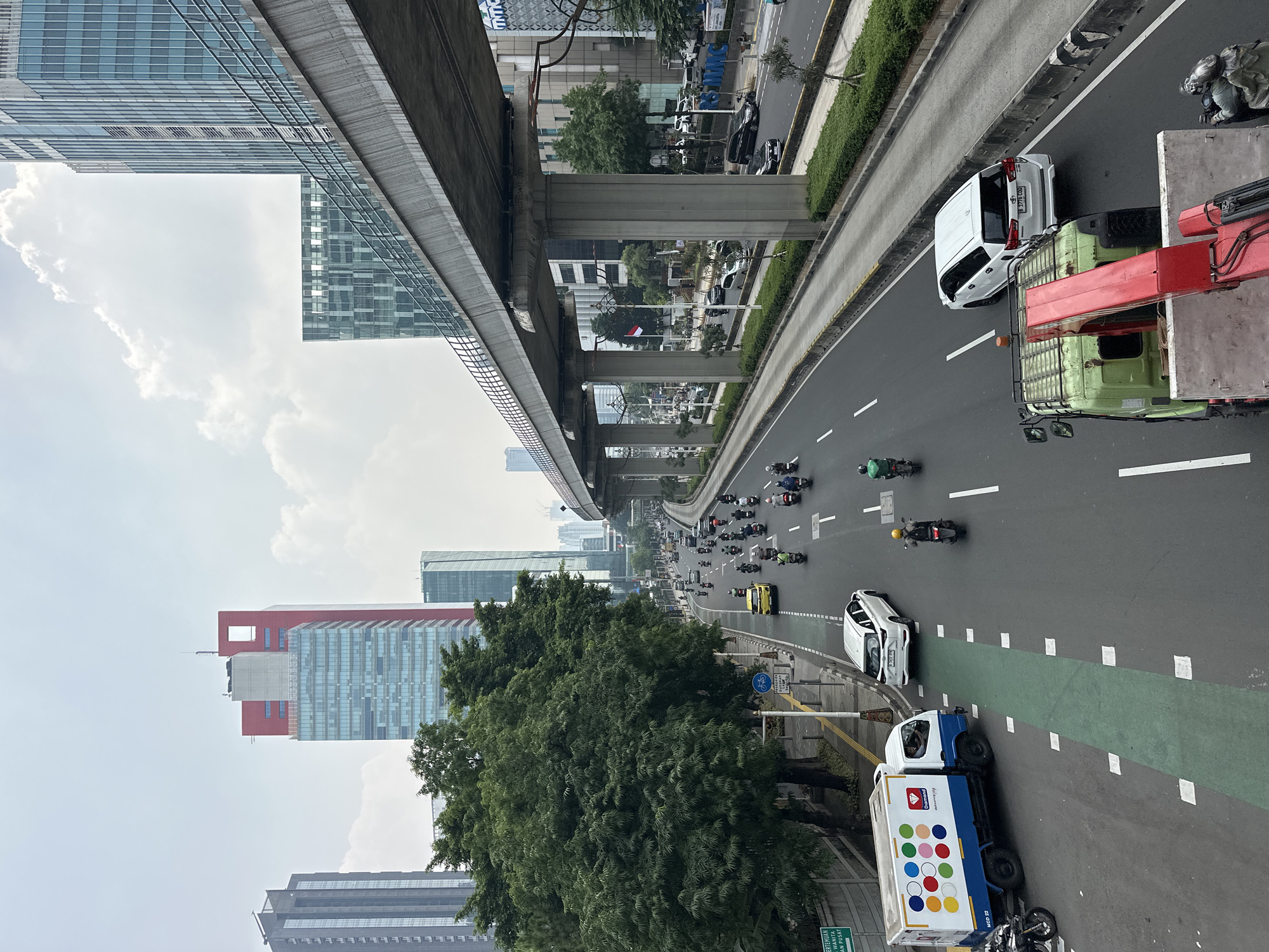 The image captures a busy street scene in South Jakarta, Indonesia, under an overcast sky. A multi-lane road is filled with vehicles, primarily motorcycles, but also cars and a truck. On the left side of the road, there is a sidewalk with trees and signage, and beyond that, several tall buildings, including a distinctive red and glass skyscraper. A raised railway or monorail track runs overhead, supported by concrete pillars. The road itself has clear lane markings, including a dedicated green lane for bicycles or motorcycles, and a white truck with colorful circles on its side is parked on the left. Further down the road, a white SUV and other vehicles are in motion. On the right side of the road, more vehicles are visible, along with a median strip of green foliage and more buildings in the background. The overall impression is one of urban activity and infrastructure.
