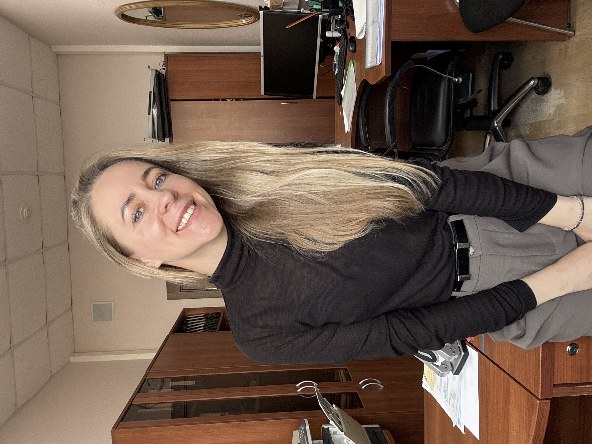 This image captures a smiling woman in an office setting in Chaikovsky, Russia. The woman, who has long, straight blonde hair and blue eyes, is looking directly at the camera with a bright, open-mouthed smile. She is wearing a black long-sleeved turtleneck shirt and grey trousers, secured with a black belt. A dark-colored beaded bracelet is visible on her left wrist, which rests casually in the foreground.
The background shows a typical office environment. Behind the woman is a wooden desk with a black computer monitor, which appears to be off, and various papers and office supplies like a stapler. To her right, a black office chair is partially visible. Further back, there are wooden cabinets or shelving units, suggesting storage or display. The walls are a neutral beige color, and a grid of white ceiling tiles with a square vent is visible in the upper left. A mirror with an oval, possibly gold or wooden, frame is mounted on the wall above the desk. The overall lighting suggests it is daytime indoors. No discernible text is visible on any objects in the image.