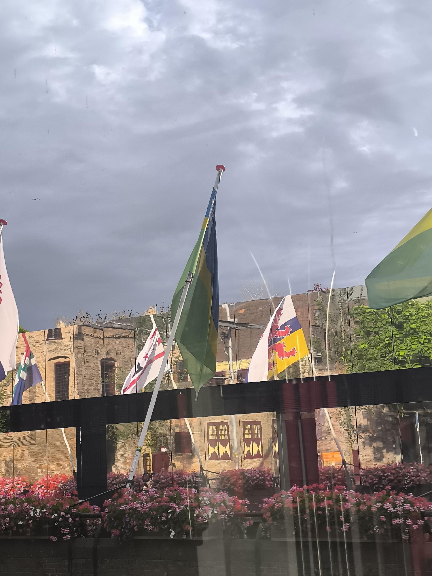 This is a vertical, eye-level, medium shot of a canal in The Hague, Netherlands, on a cloudy day. Multiple flags are visible flying from poles on a moored boat. From left to right, the flags include a white flag with red lettering, a blue, green, and white flag, a green and blue flag, and a green and yellow flag. The background features a brick building with closed shutters and windows, reflected in a mirrored surface along the canal. A railing with flower boxes filled with pink and red flowers runs along the foreground, obscuring the lower portion of the boat and canal. The sky is overcast and gray, with some lighter patches indicating breaks in the clouds. The overall impression is one of a bright, tourist-friendly setting despite the cloudy weather.