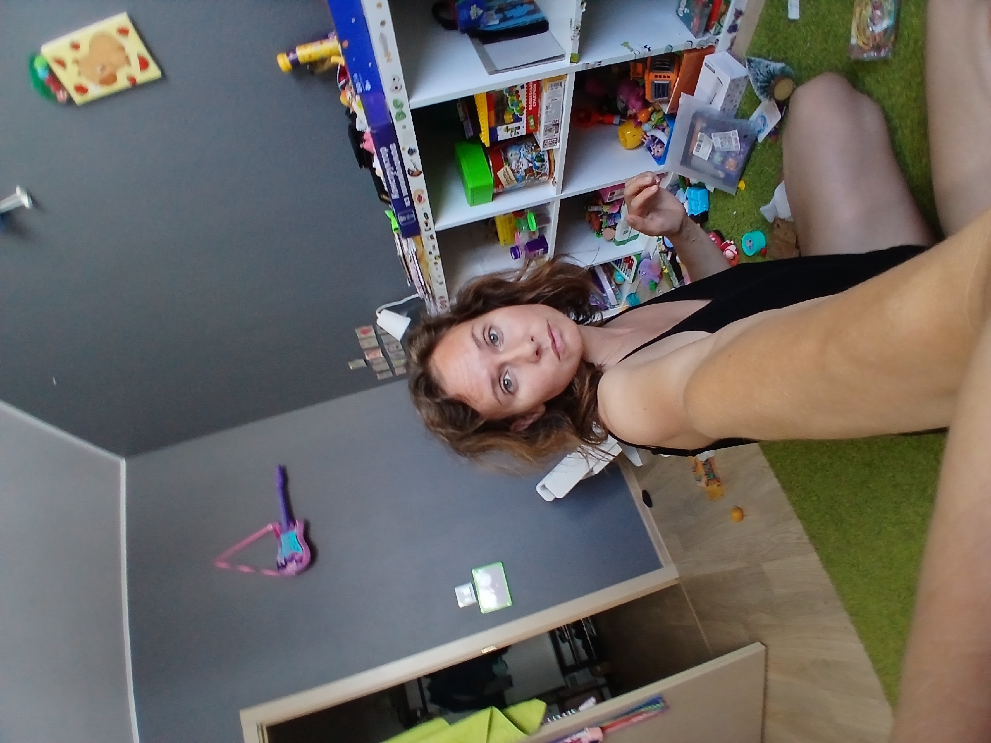 A woman is sitting on a green rug in a room that appears to be a child's playroom. The room has gray walls and a wooden floor. There is a white shelf unit filled with toys and books, a doorway to another room, and a small plastic guitar hanging on the wall. The woman is wearing a black top and her left arm is visible in the foreground. The overall impression is of a somewhat cluttered and lived-in space. There is no discernible information that would allow for location identification in Yekaterinburg, Russia.