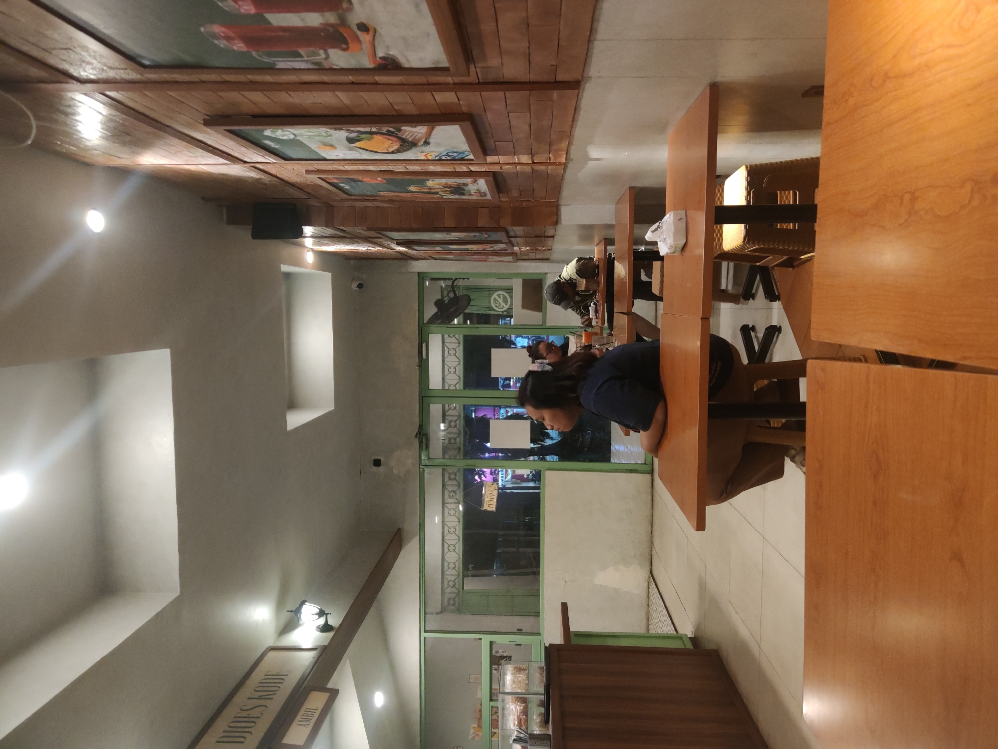 The image shows the interior of a cafe, likely in Surabaya, Indonesia, with wooden tables and chairs. In the foreground, two large wooden tables are visible. In the mid-ground, a woman in a dark blue shirt and tan pants is sitting at a table, looking to her right. She appears to be alone. Further back, two people are seated at another table. One person is wearing a cap and appears to be looking down at their phone or food, while the other person's face is not fully visible. The cafe has brick-patterned walls adorned with framed pictures. The overall lighting is dim, with bright overhead lights casting a warm glow. The cafe appears to be a casual dining establishment.